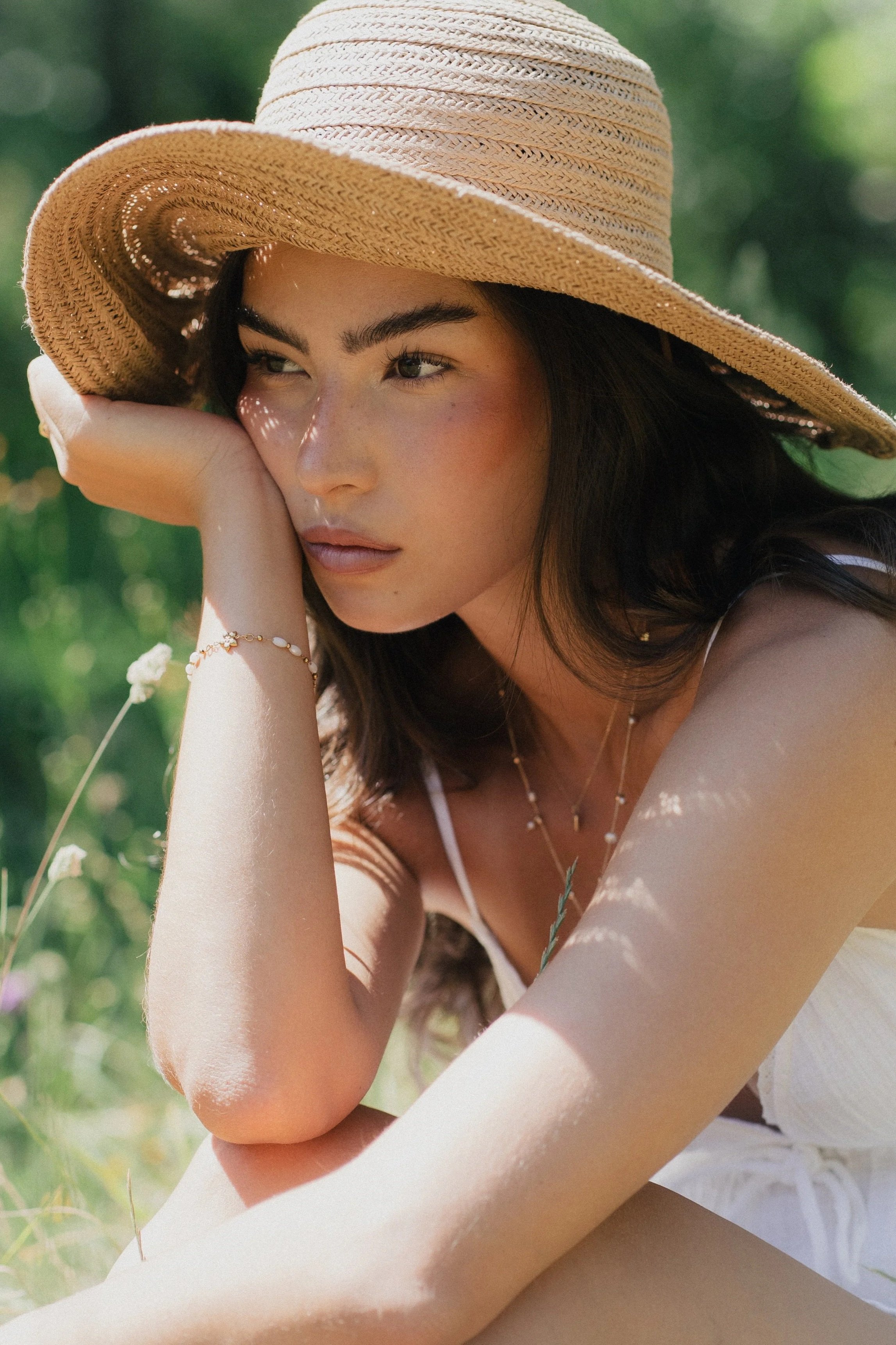 A young woman with dark hair and light makeup wearing a large straw sunhat and jewelry, resting her chin on her hand outdoors in a sunny, green environment.