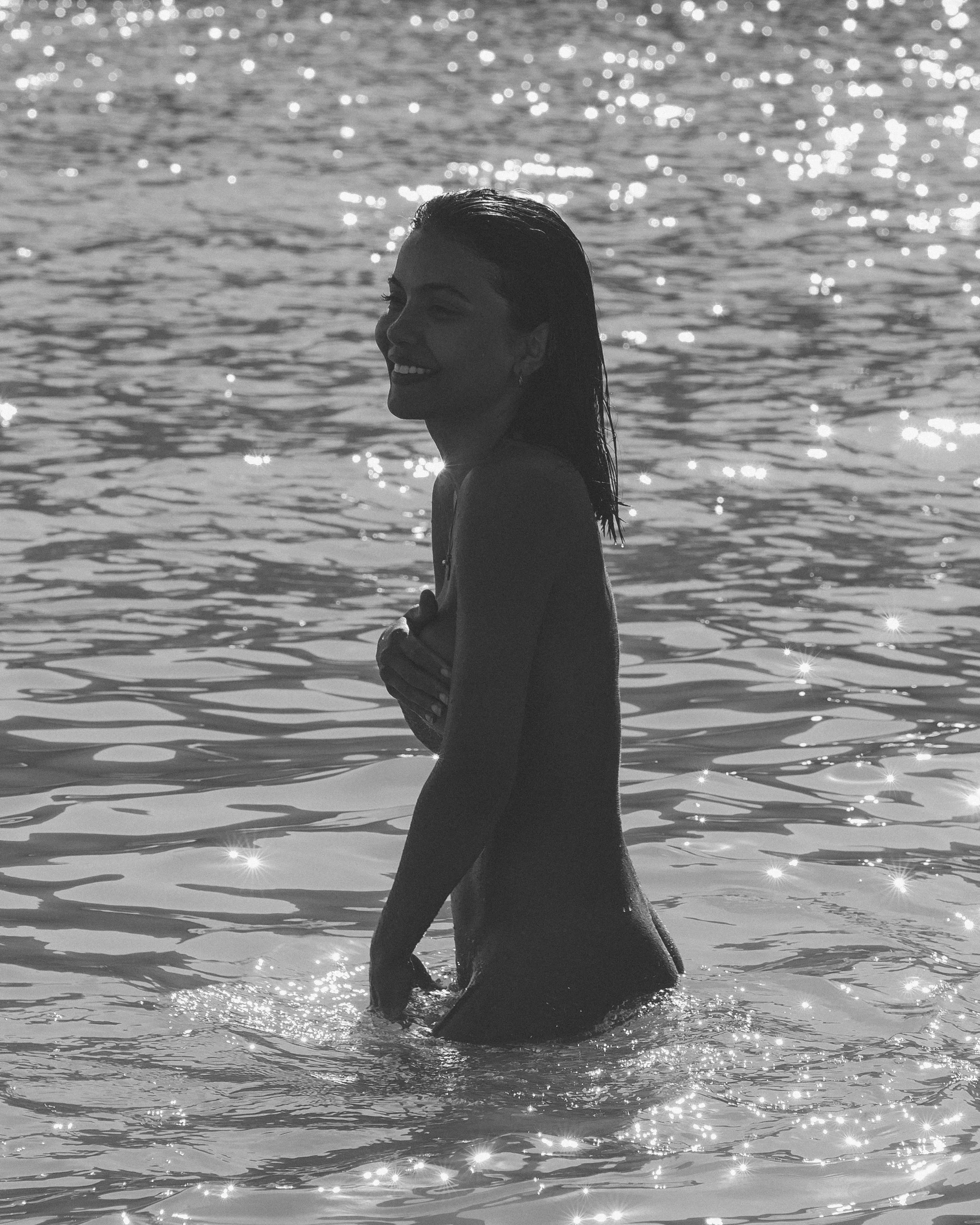 A woman with wet hair smiling and holding her hands to her chest while standing waist-deep in a body of water, with sunlight reflecting on the surface.