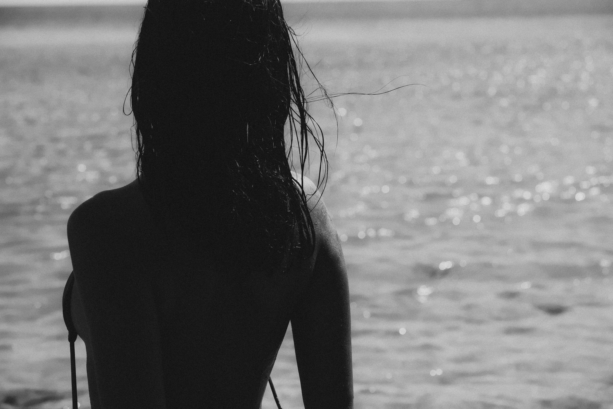 Back view of a woman with long wet hair sitting at the beach, facing the water in black and white.
