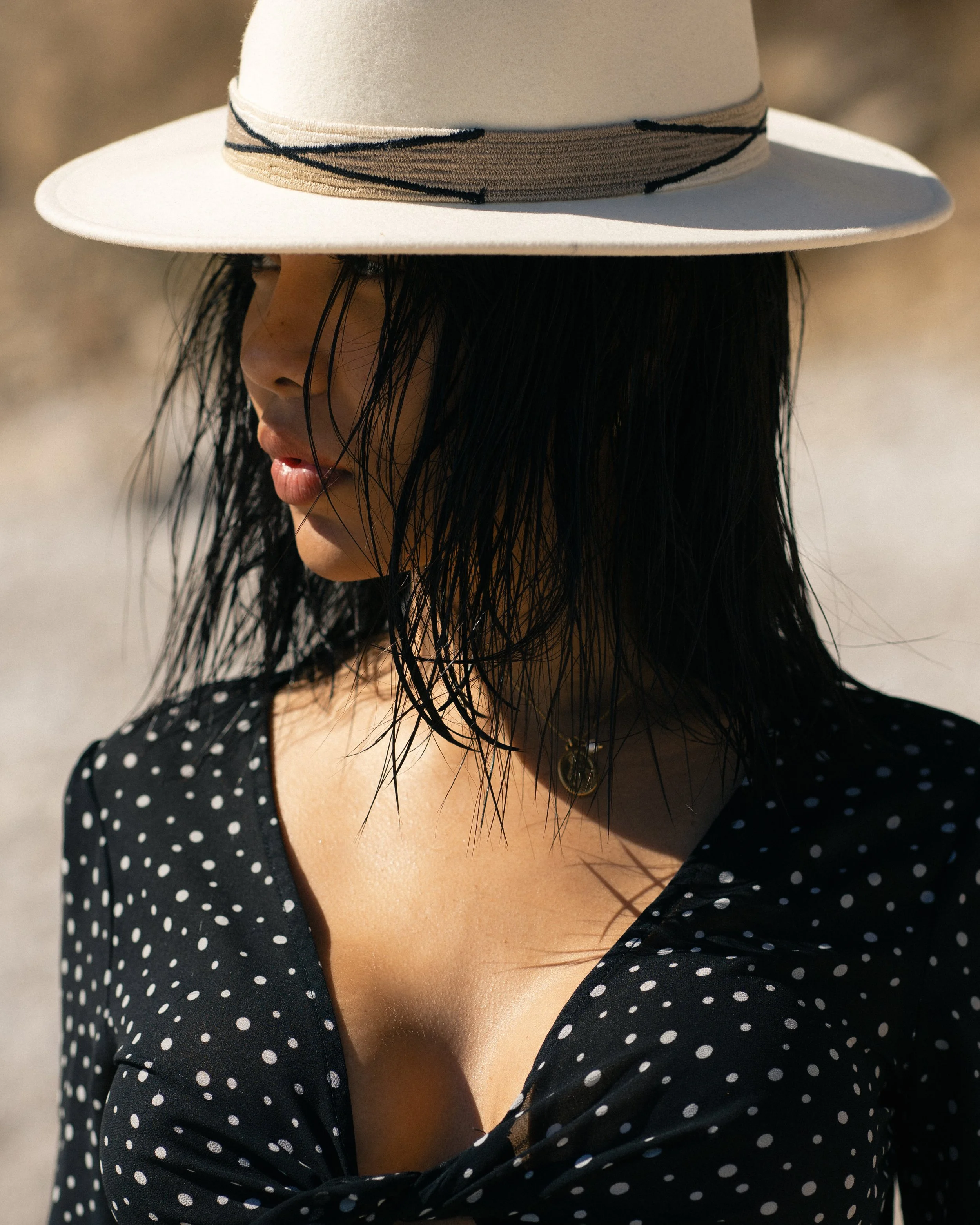 A woman with long dark wet hair wears a wide-brimmed beige hat with black trim, a black dress with white polka dots, and a gold necklace, standing outdoors in blurred natural background.
