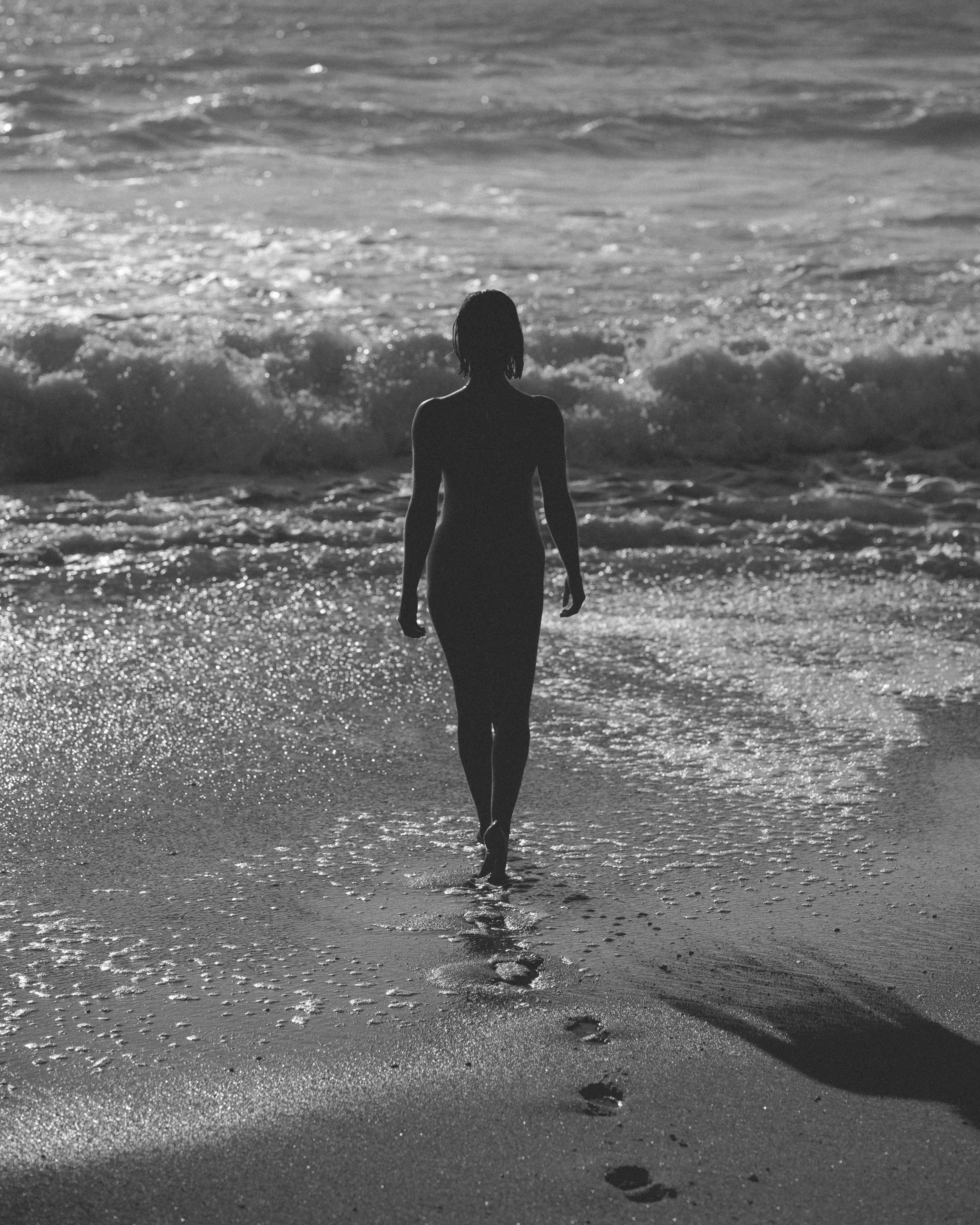 Silhouette of a woman walking along the shoreline at sunset or sunrise, with water and waves in the background and footprints in the sand.