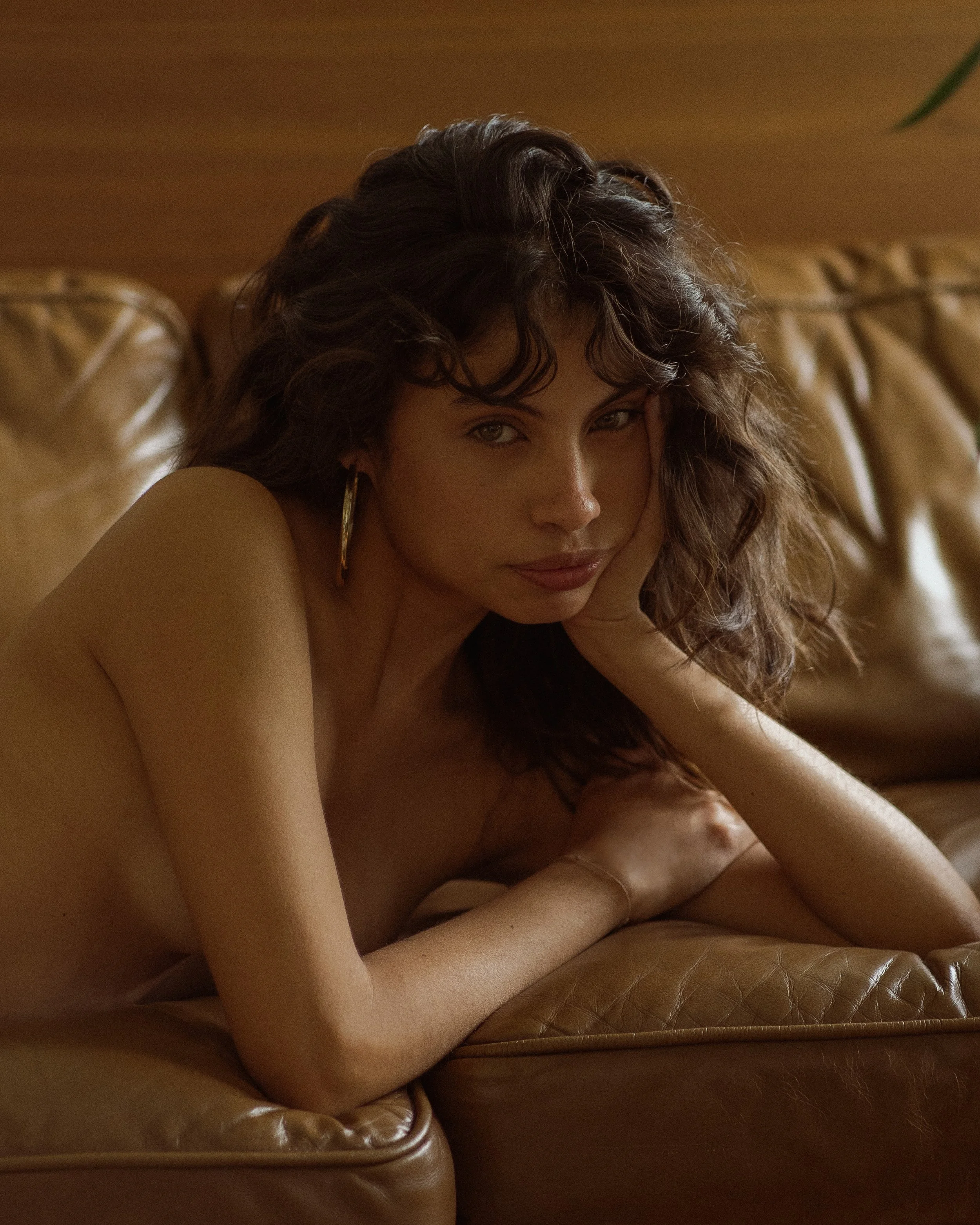 A woman with curly brown hair and hoop earrings resting her head on her hand, sitting on a leather couch with warm lighting.