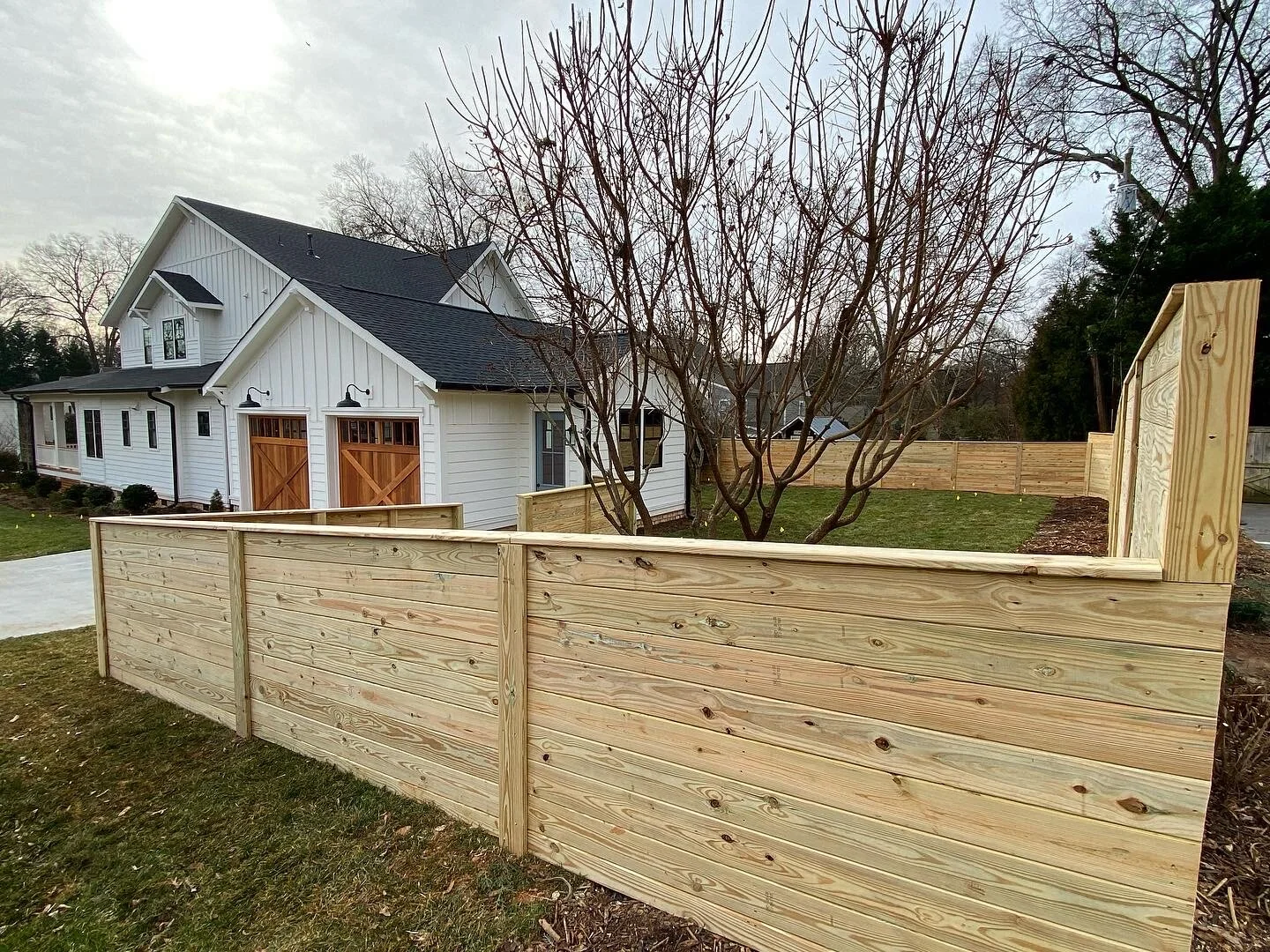 A backyard with a newly built wooden fence surrounding a small grassy area and a leafless tree. In the background, there is a white two-story house with black roofing, garage doors, and outdoor lighting. The sky is overcast with the sun partially obscured.