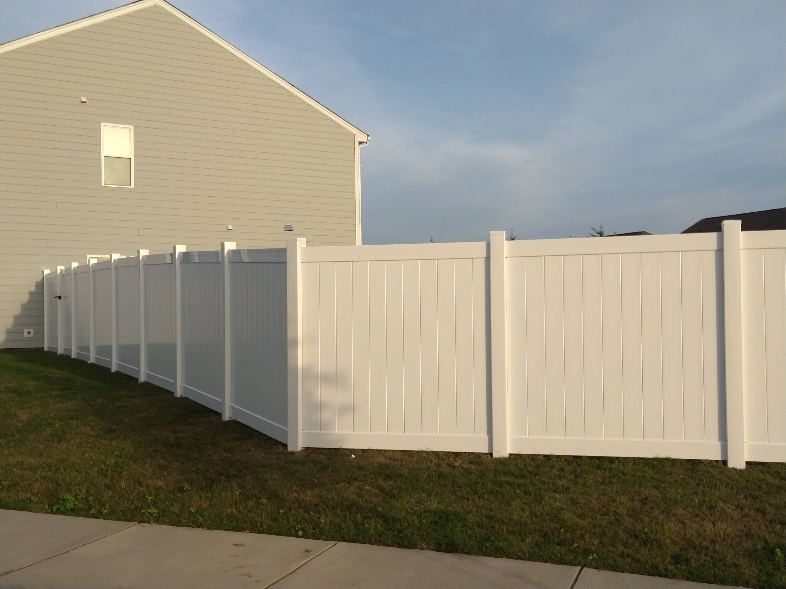 A white vinyl privacy fence surrounds a grassy backyard next to a beige house with a window, under a partly cloudy sky.