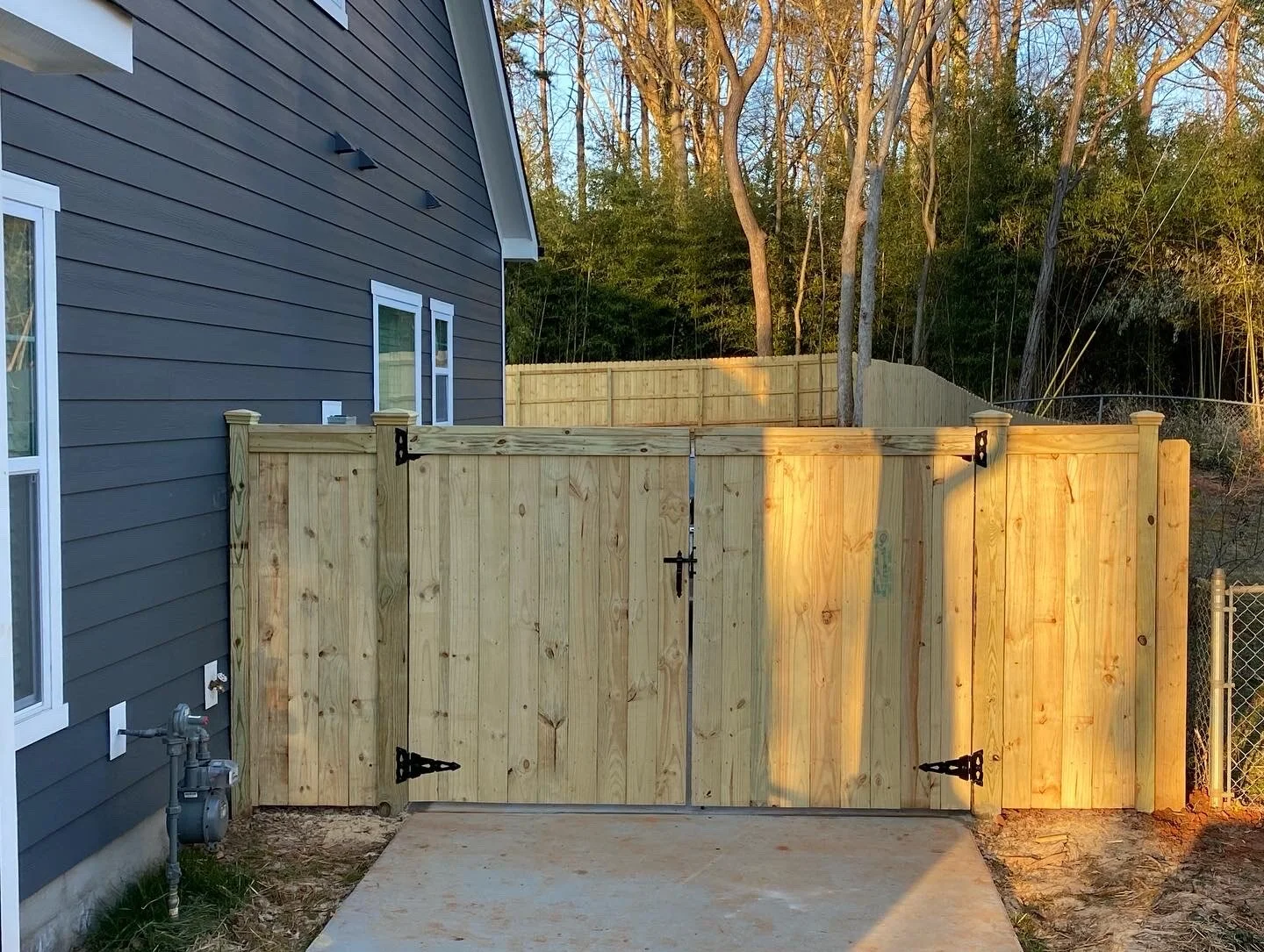 New wooden fence gate with black hinges and latch in front of a dark blue house with white window trim, next to a concrete patio, with trees and a wooden privacy fence in the background.