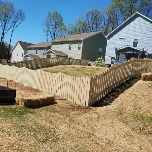A backyard with a wooden fence, some grass, and neighboring houses under a clear blue sky.