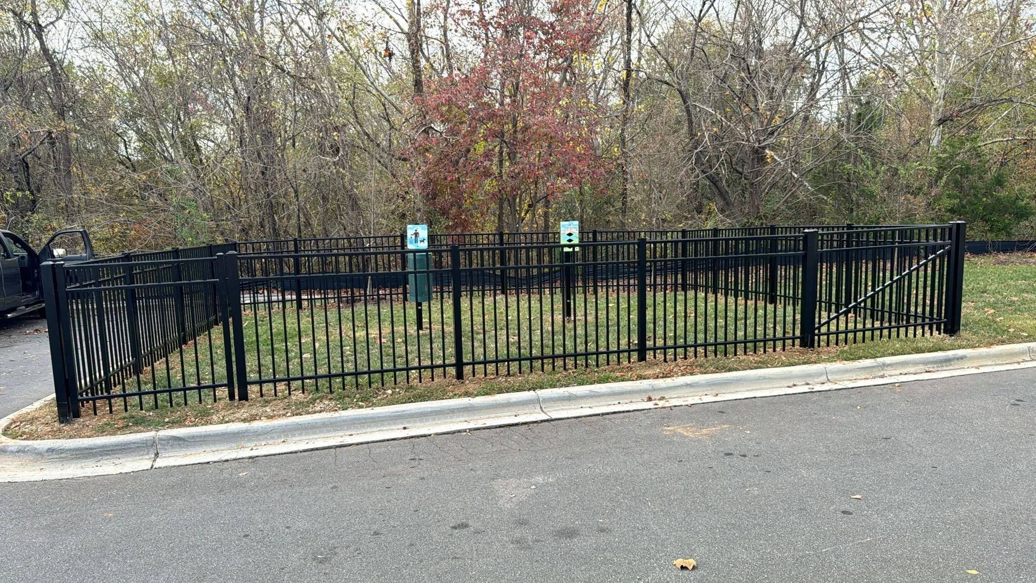 A small fenced area in a park with two signs, one depicting a person and a dog, and the other with a fish symbol. Trees and a grassy area are behind the fence.