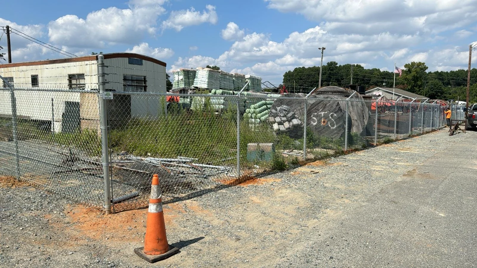 Construction yard with construction materials, equipment, and a chain-link fence surrounding the area, under partly cloudy sky.