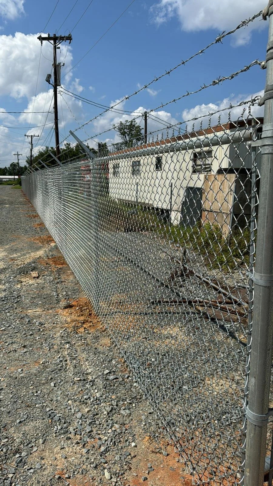 A chain-link fence with barbed wire on top, running alongside a gravel pathway in an outdoor area. In the background, there are utility poles, a white mobile home, and a blue sky with some clouds.