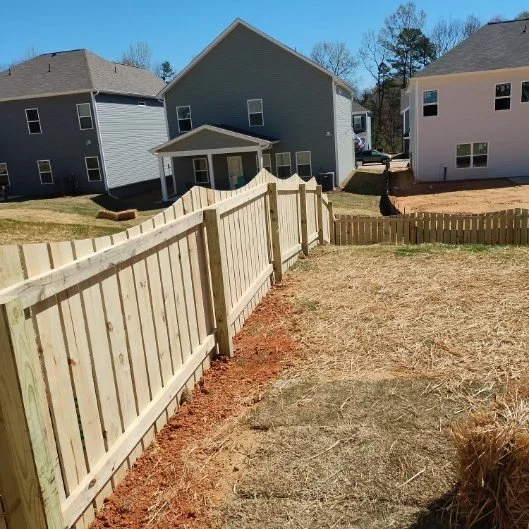 New wooden fence partially installed in a backyard with houses in the background during daytime.
