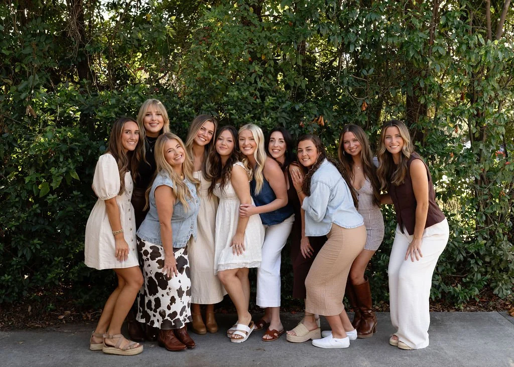 Ten women standing together outdoors, smiling in front of green bushes, dressed in casual summer outfits.