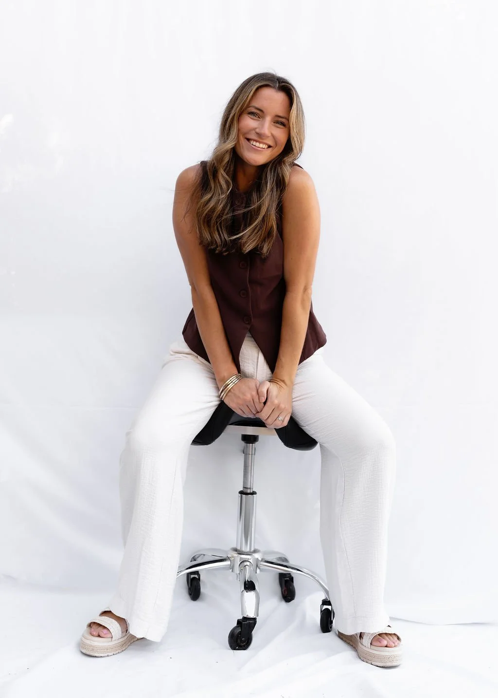 A woman sitting on a black office stool against a plain white background, smiling, dressed in a sleeveless brown top, white pants, and beige platform sandals, with bracelets on her left wrist.