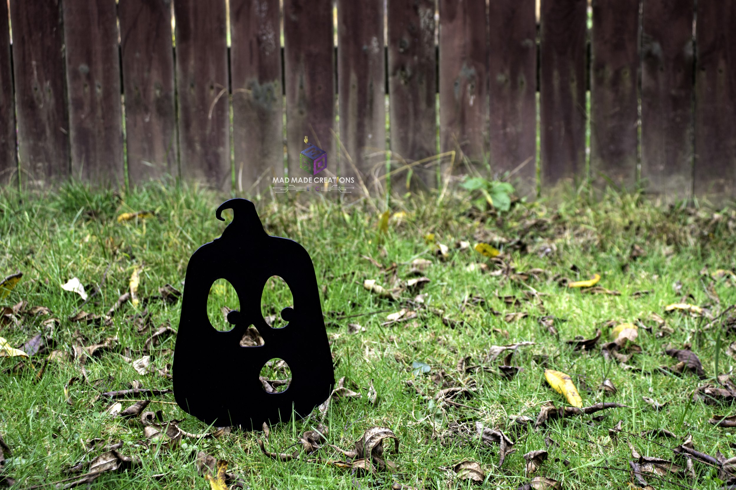 Close-up view of black 3D printed OH pumpkin yard stake by MAD Made Creations, placed on autumn grass in front of wooden fence – Halloween outdoor lawn decoration