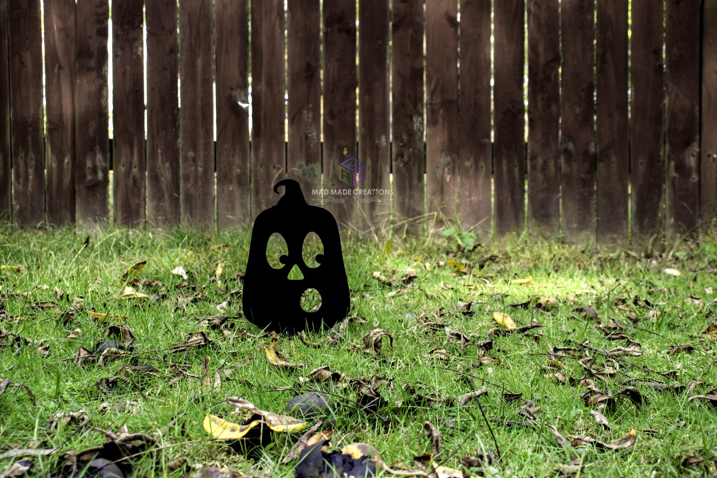3D printed Halloween OH pumpkin yard stake with spooky face design by MAD Made Creations, displayed on fall grass in front of a wooden fence during daytime