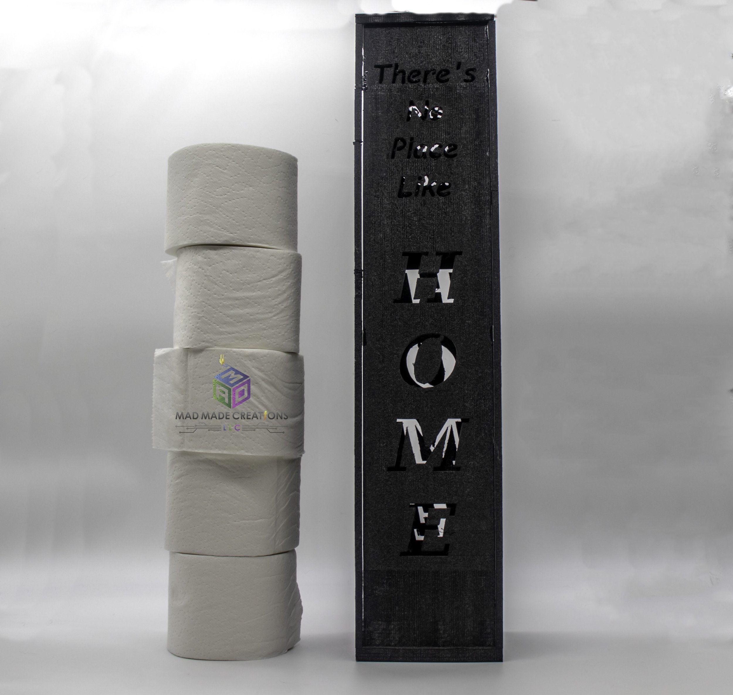 Black custom 3D printed toilet paper holder with vertical cutout text 'There's No Place Like HOME' next to a stack of white toilet paper rolls – decorative bathroom storage by MAD Made Creations