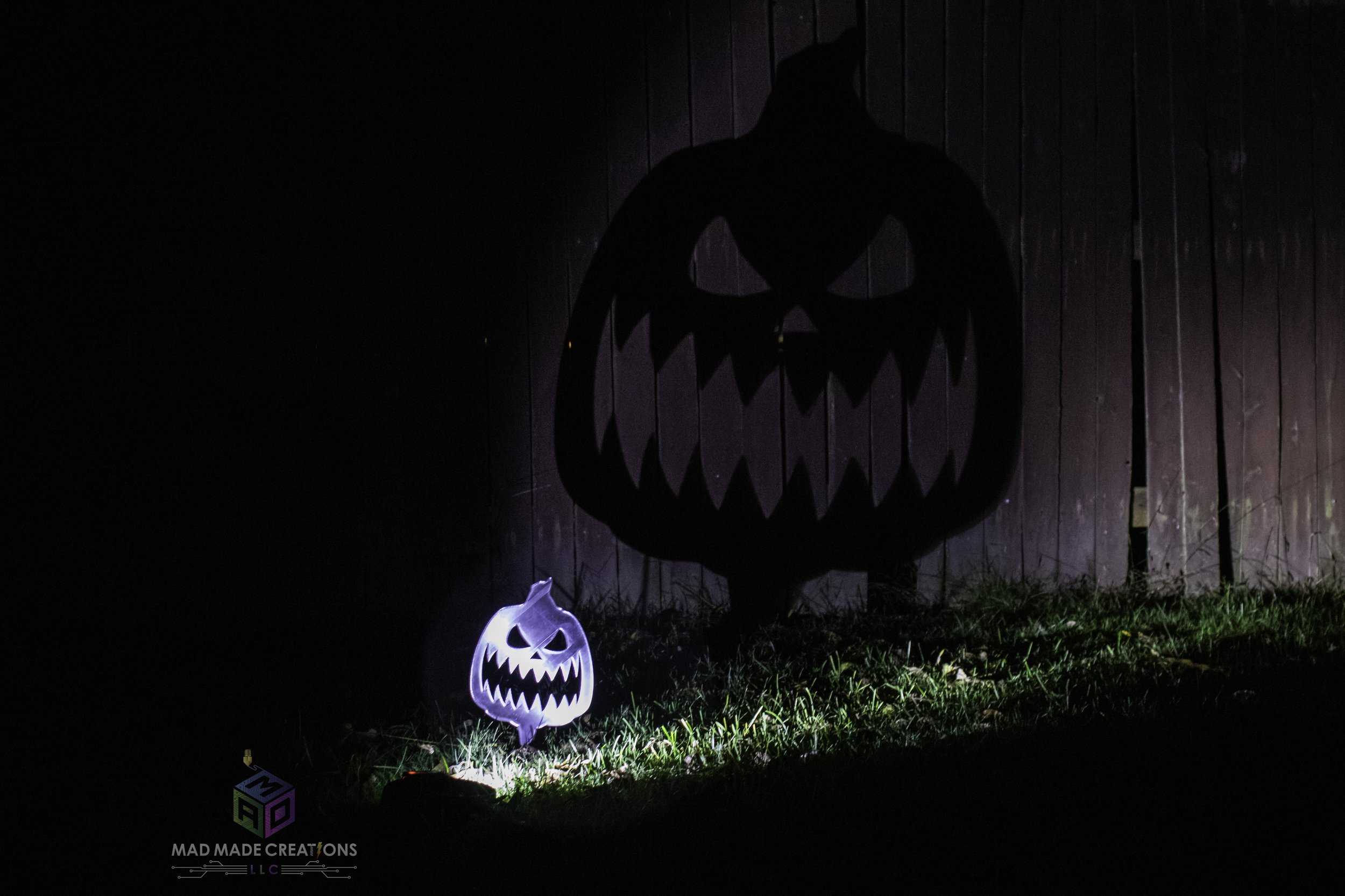 Illuminated 3D printed scary pumpkin yard stake casting a large menacing jack-o'-lantern shadow on a wooden fence at night, handmade Halloween decoration by MAD Made Creations