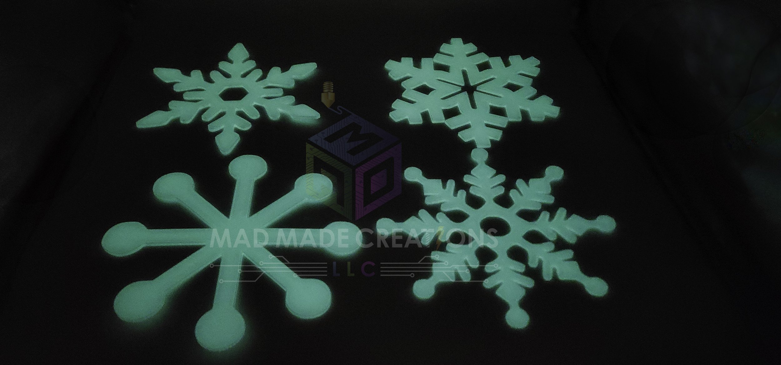 Pack of four 12-inch hangable 3D-printed glow-in-the-dark white snowflake ornaments glowing teal against a dark background