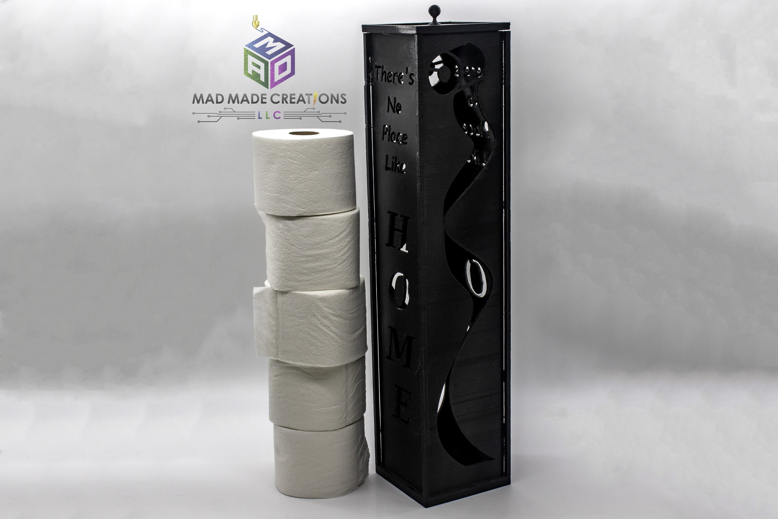 Vertical black 3D printed toilet paper holder with 'There's No Place Like HOME' cutout design, standing next to stacked toilet paper rolls – decorative bathroom storage by MAD Made Creations