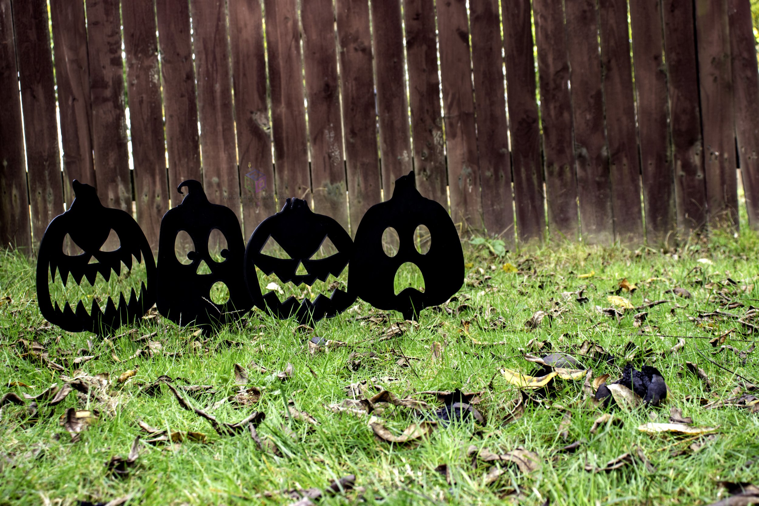 Set of four 3D printed black Halloween pumpkin yard stakes with unique spooky faces, displayed on grass in front of a rustic wood fence, handmade by MAD Made Creations