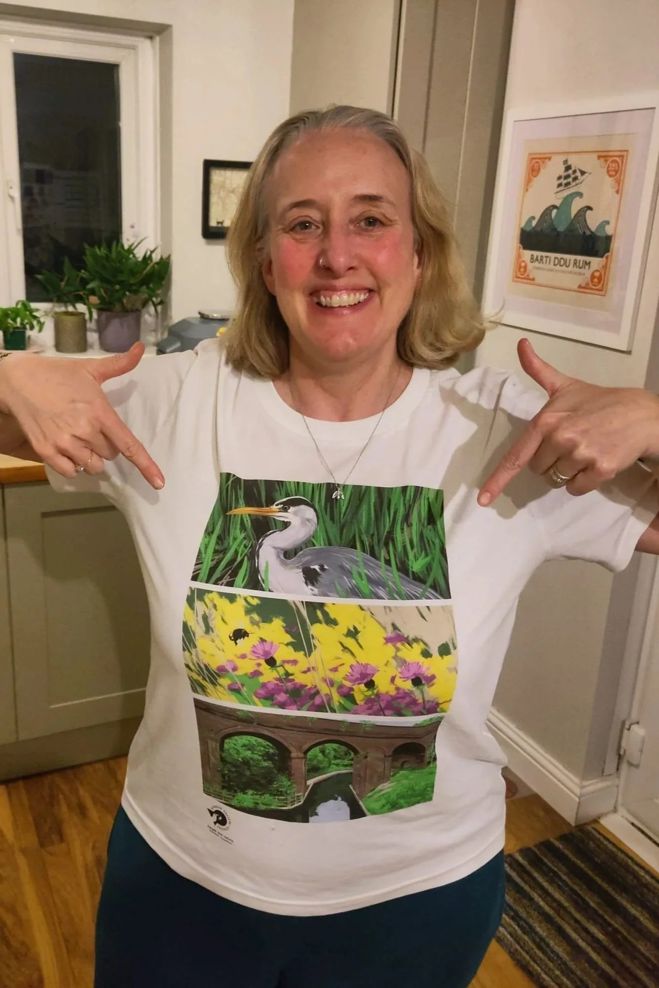 Staff member Sharon is wearing one of SVP's T shirts with a heron at the top, then a bee, then the arches of Capels Mill. She is pointing to the design and smiling.