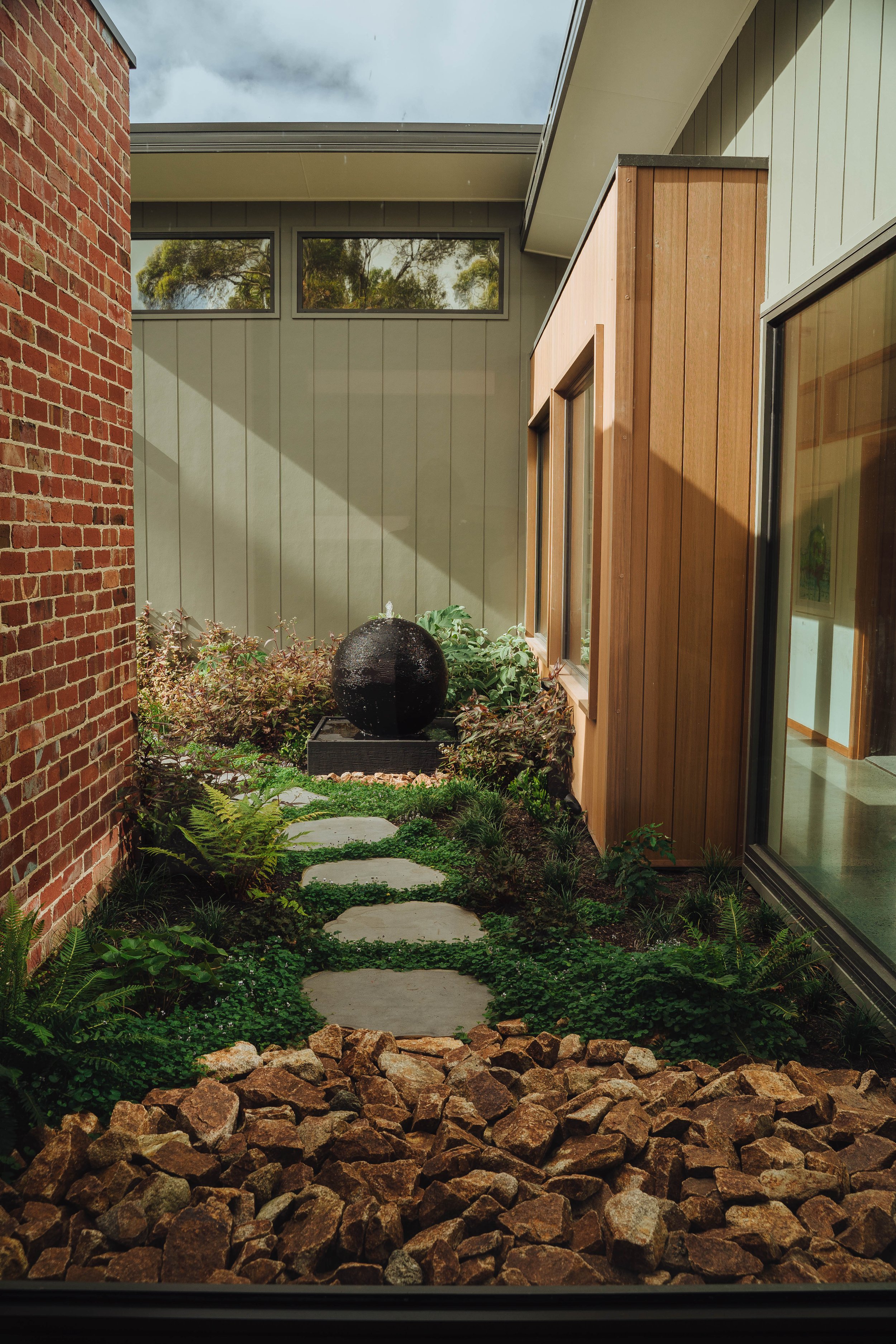 A small landscaped garden with stepping stones, plants, rocks, and a black spherical water fountain between a brick wall and a beige house.