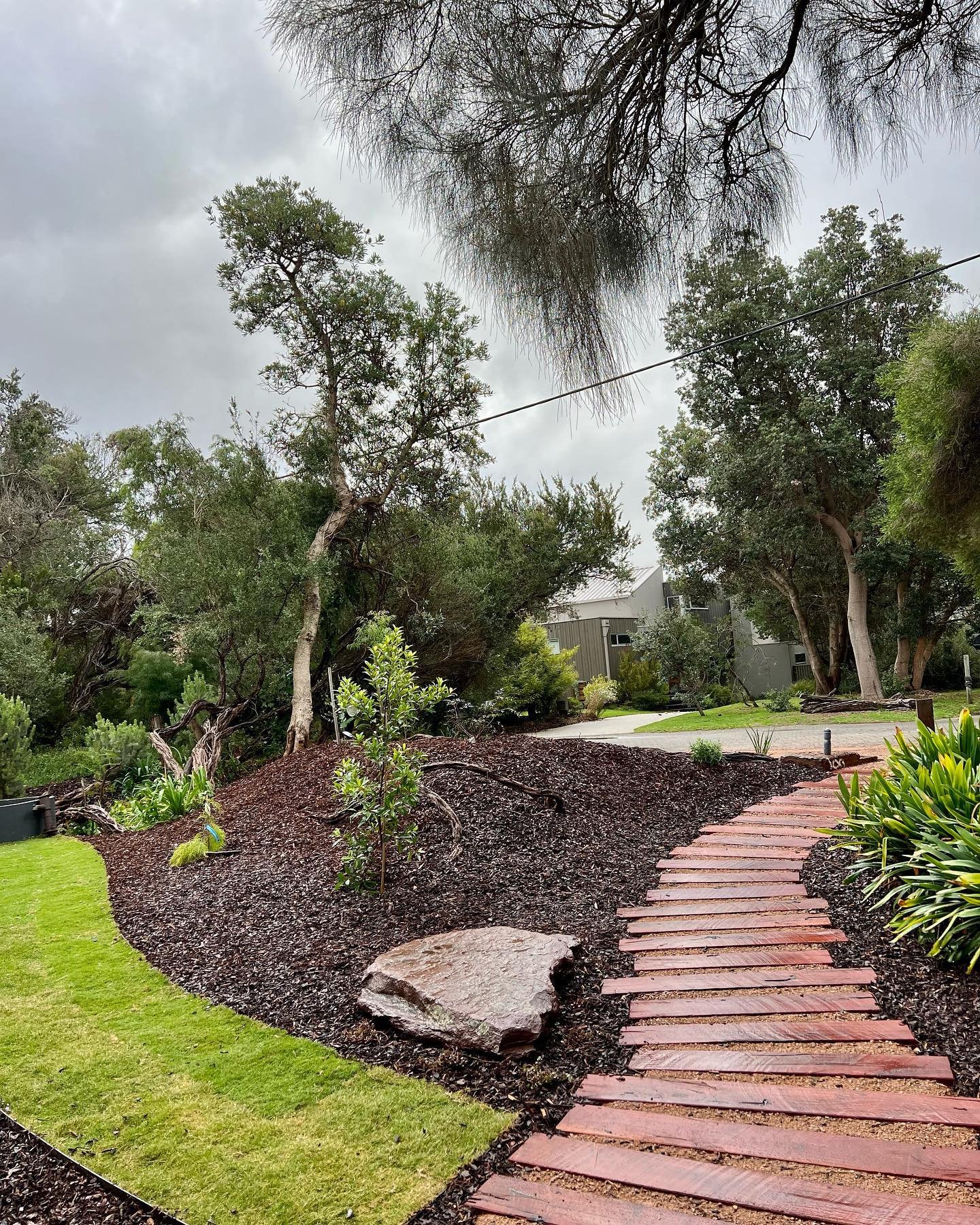 Curved brick pathway through a well-maintained garden with lush green grass, small trees, shrubs, and a cloudy sky overhead.