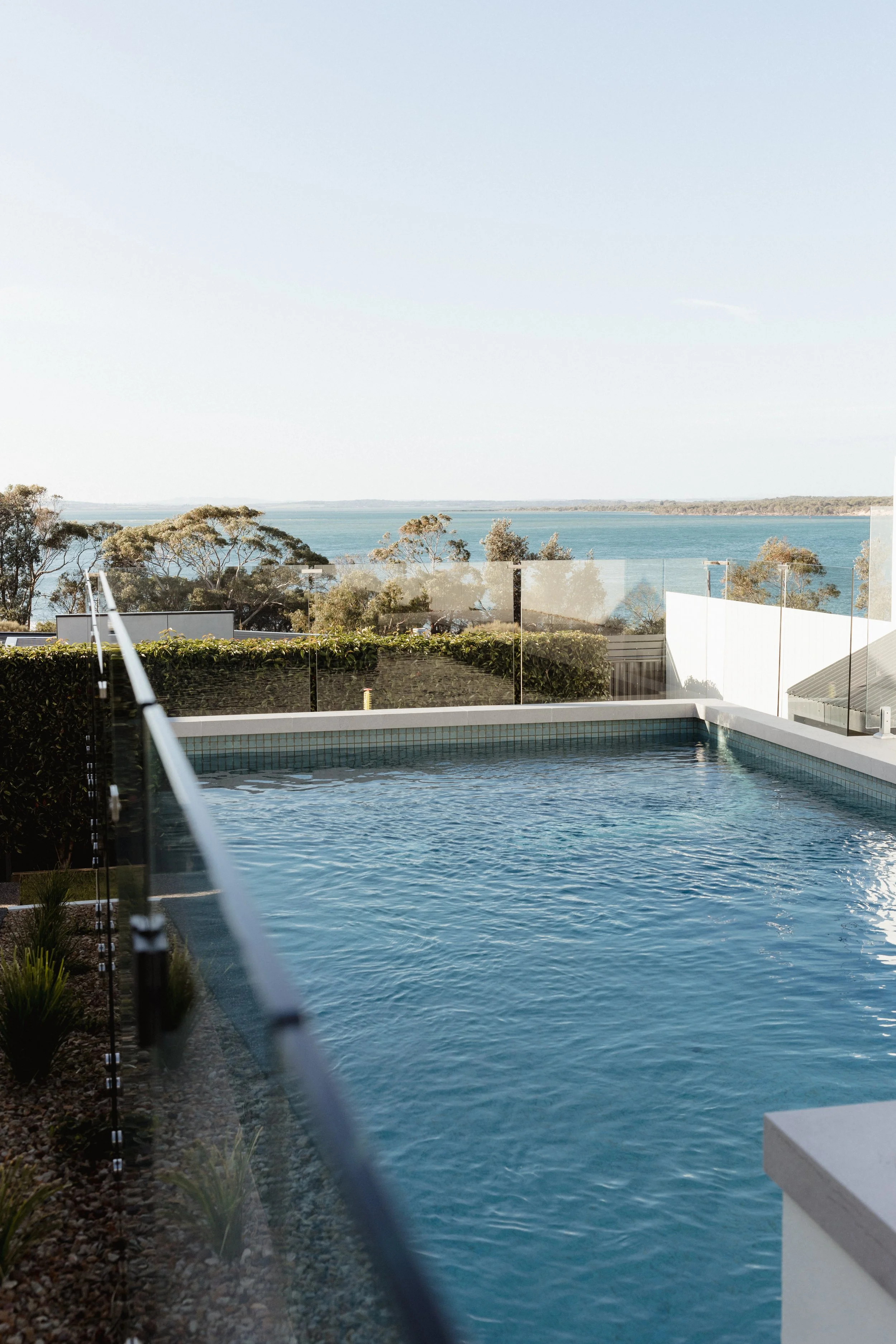 Outdoor swimming pool with a glass safety barrier, overlooking trees, water, and horizon in the distance under a clear sky.