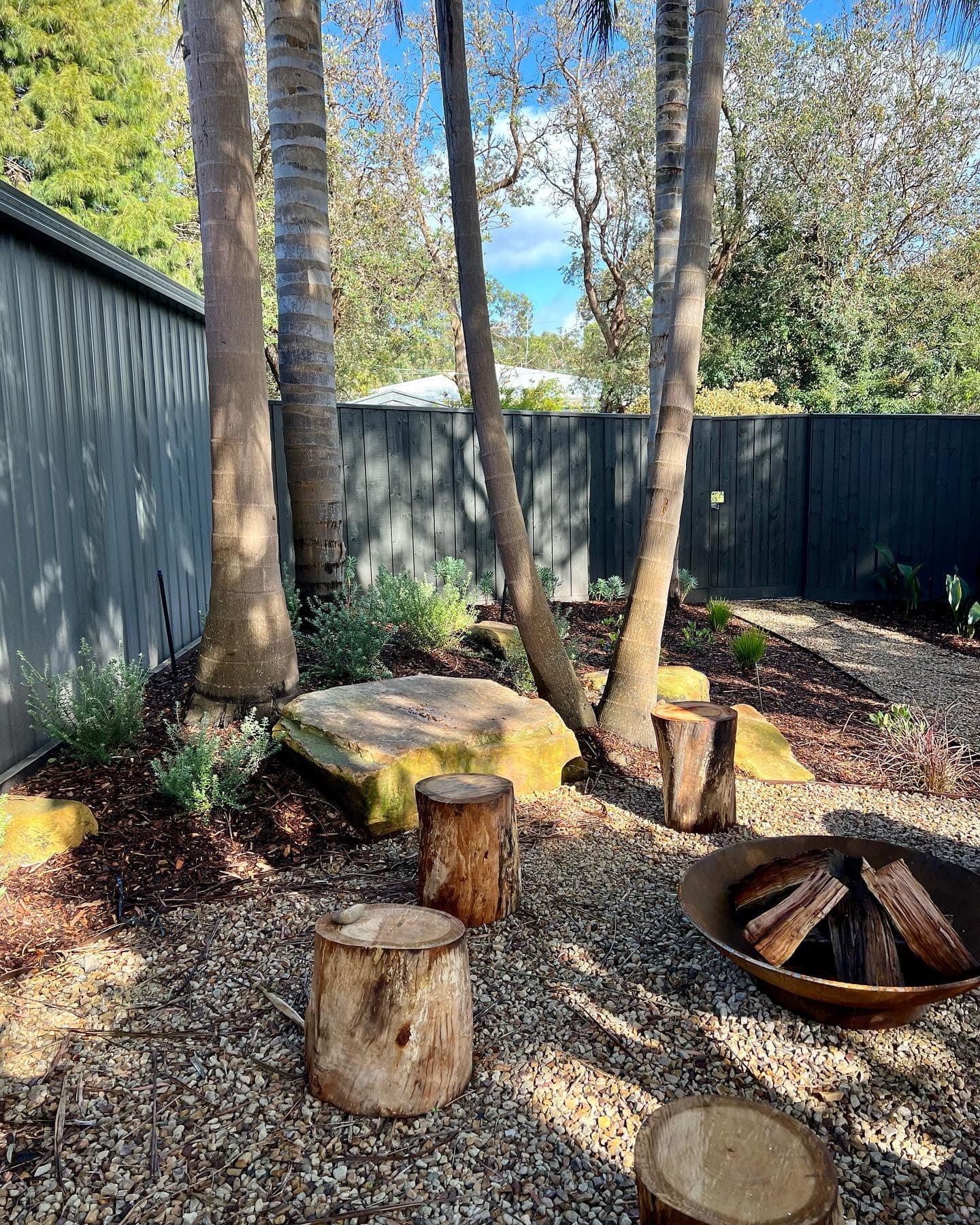Backyard with a black wooden fence, three palm trees, shrubbery, large rocks, and a firepit filled with split logs, all on a bed of small pebbles.