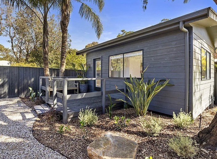 Gray backyard shed with a small deck, surrounded by plants, trees, and a gravel pathway.