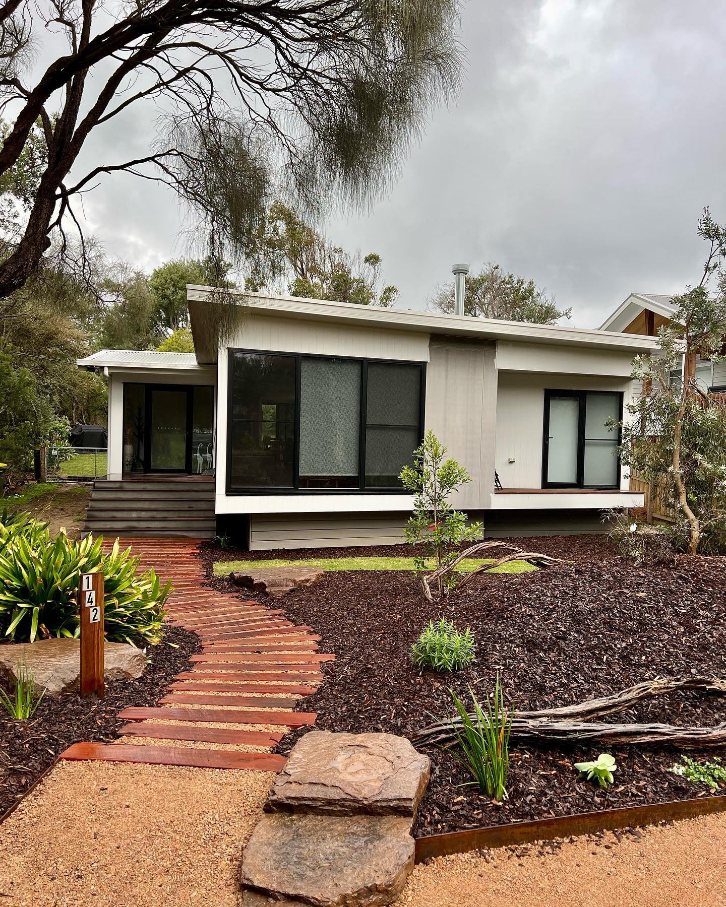 Modern white house with black window frames and sliding glass doors, surrounded by a landscaped yard with a curved brick pathway, plants, and trees, under a cloudy sky.