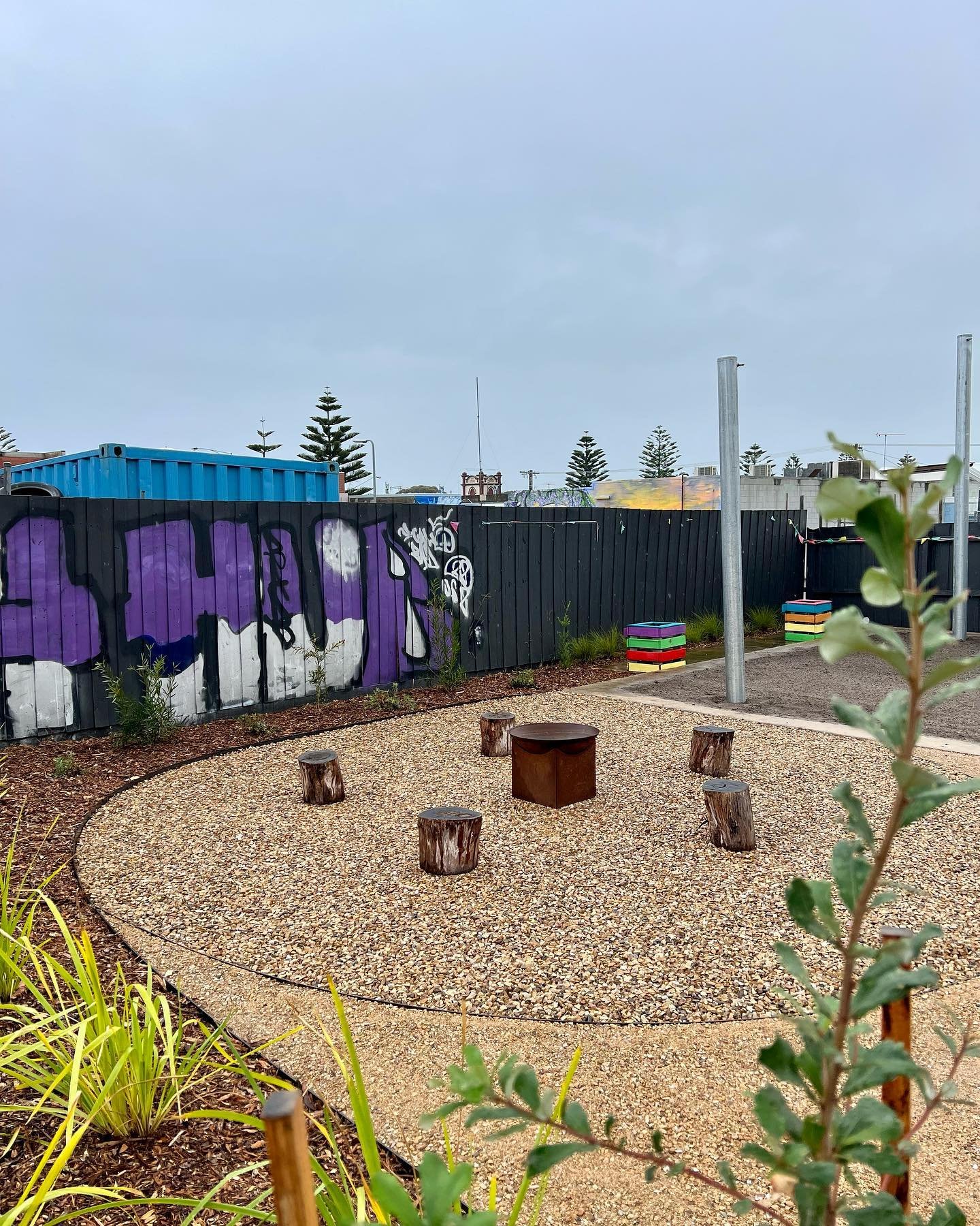 A small outdoor seating area with tree stumps arranged in a circle around a metal fire pit, set on gravel, with a black fence, colorful painted block structures, and graffiti in the background.