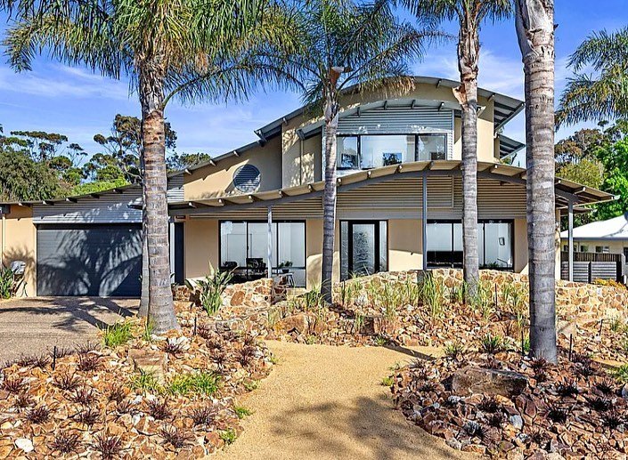 Modern two-story house with large windows, surrounded by palm trees and a landscaped yard with rocks and drought-tolerant plants.