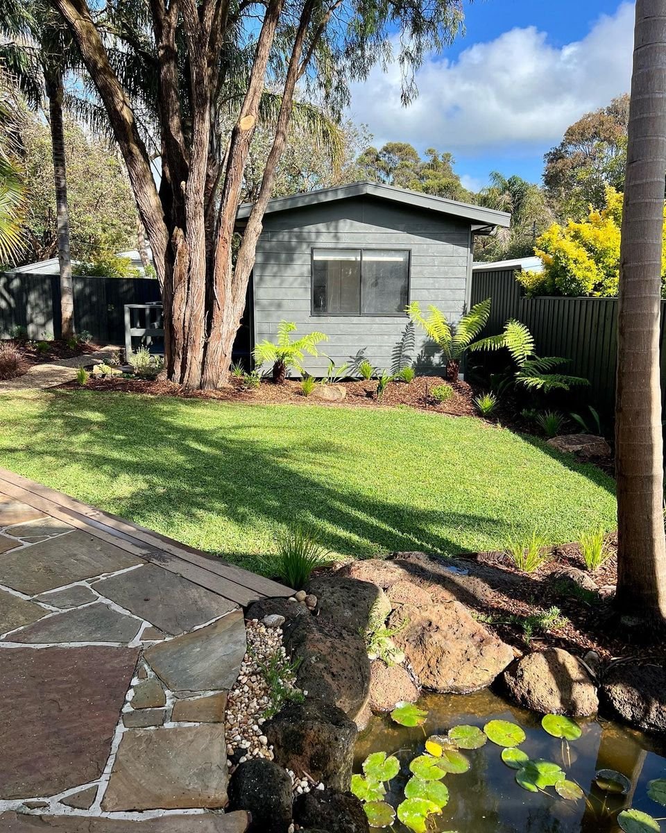 A small gray house with a sloped roof, surrounded by lush green grass, trees, and tropical plants. A stone pathway is visible in the foreground near a pond with lily pads.