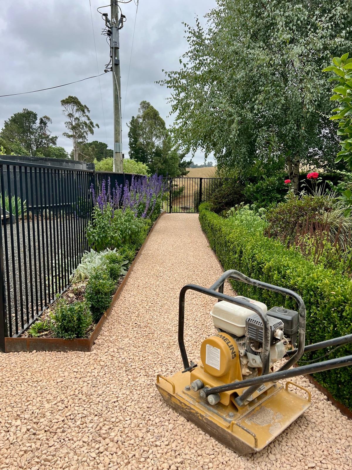 A gravel pathway bordered by green bushes and flowers on either side, with a black metal fence and a yard with flowering plants on the sides, leads to a black gate in the distance. There is a portable plate compactor on the pathway in the foreground,