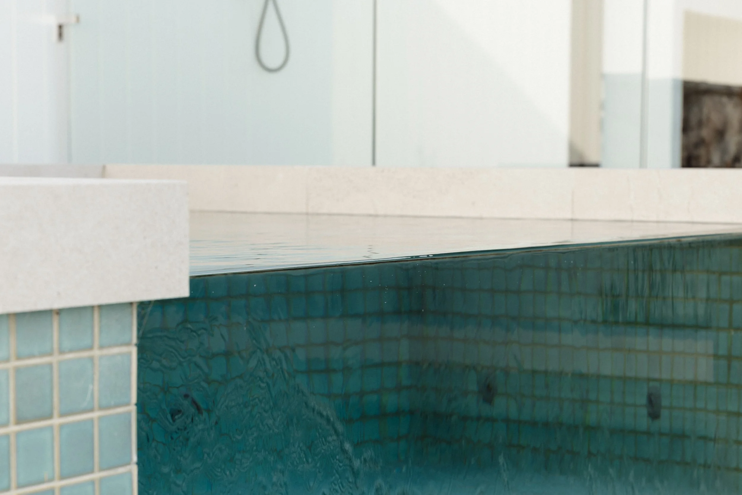 Part of an indoor swimming pool with turquoise tiles and calm water. A white tiled ledge and a background with a showerhead and door are visible.