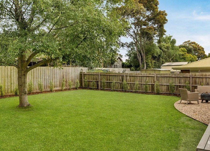 A backyard with a lush green lawn, a large shade tree on the left, a wooden fence around the yard, and a sitting area with chairs and a fire pit on the right.