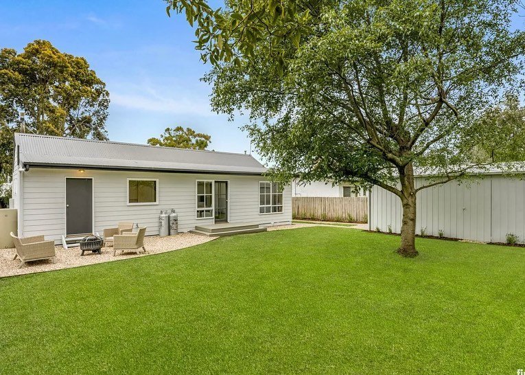 Backyard with green lawn, tree, small white house with gray roof, outdoor seating area, and a white fence.
