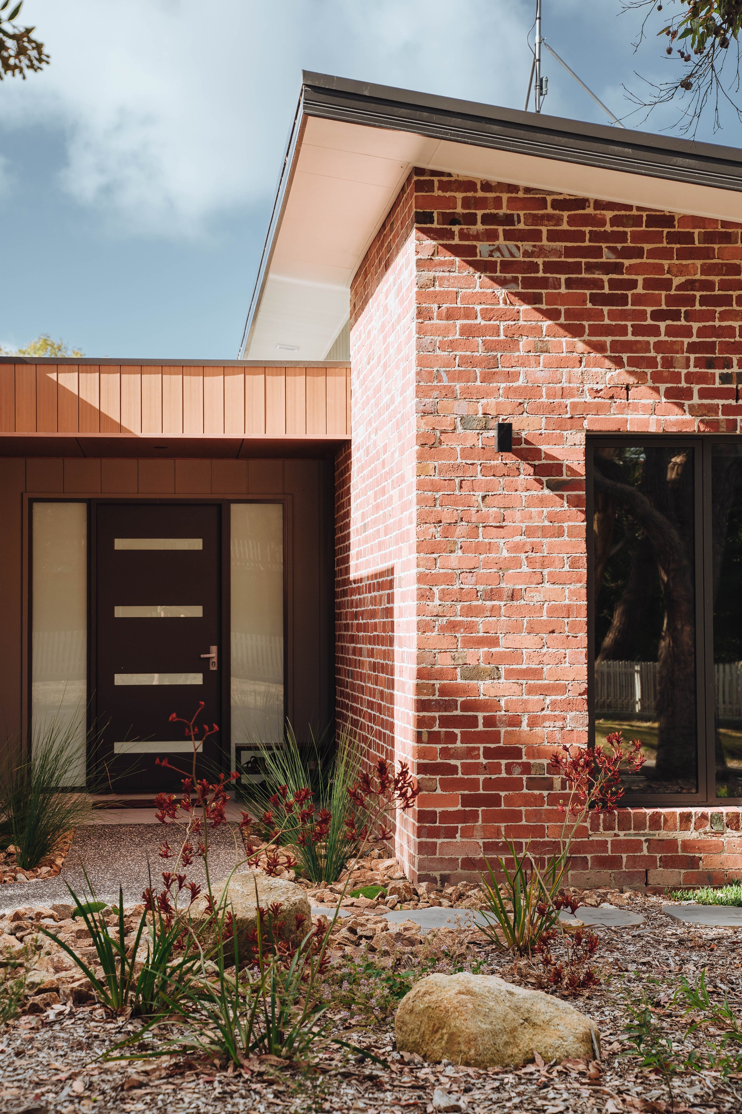 Modern house exterior with brick wall, black front door, large window, and landscaped garden with rocks and plants