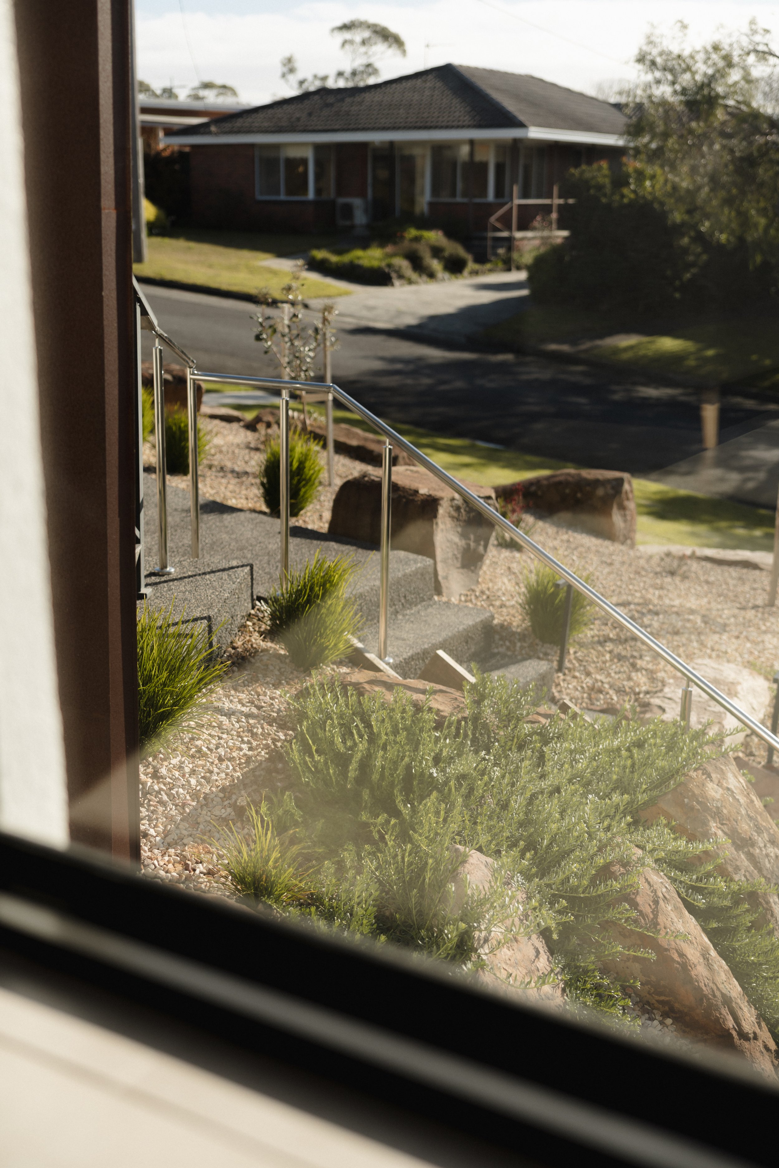 View of a suburban front yard with shrubbery, rocks, and a concrete pathway, seen through a window with a partial view of the interior wall.