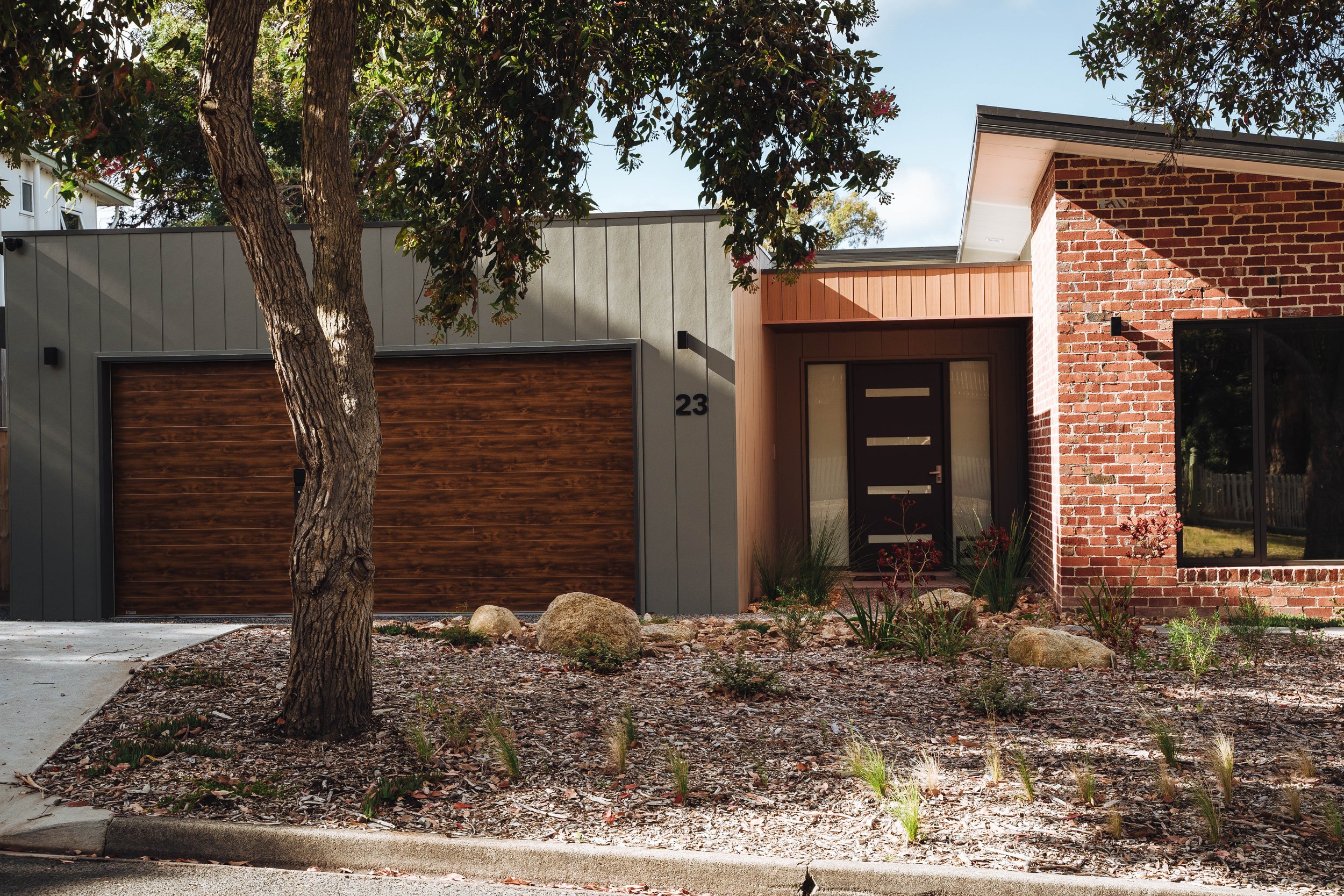 Front view of a modern house with a tree in the yard, featuring a garage with a wooden door, a brick wall, and a black front door.