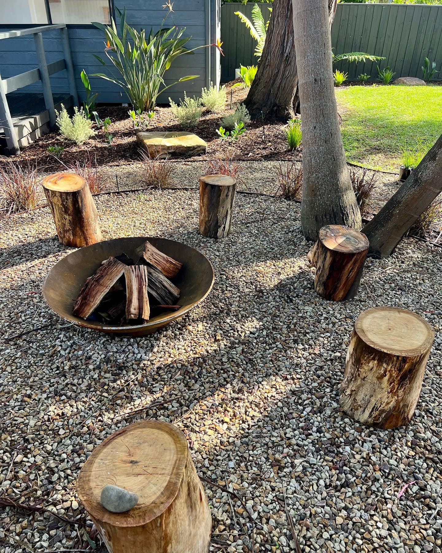 A backyard fire pit area with a metal fire bowl filled with firewood, surrounded by cut tree stumps serving as seating, on a gravel surface with plants and trees in the background.