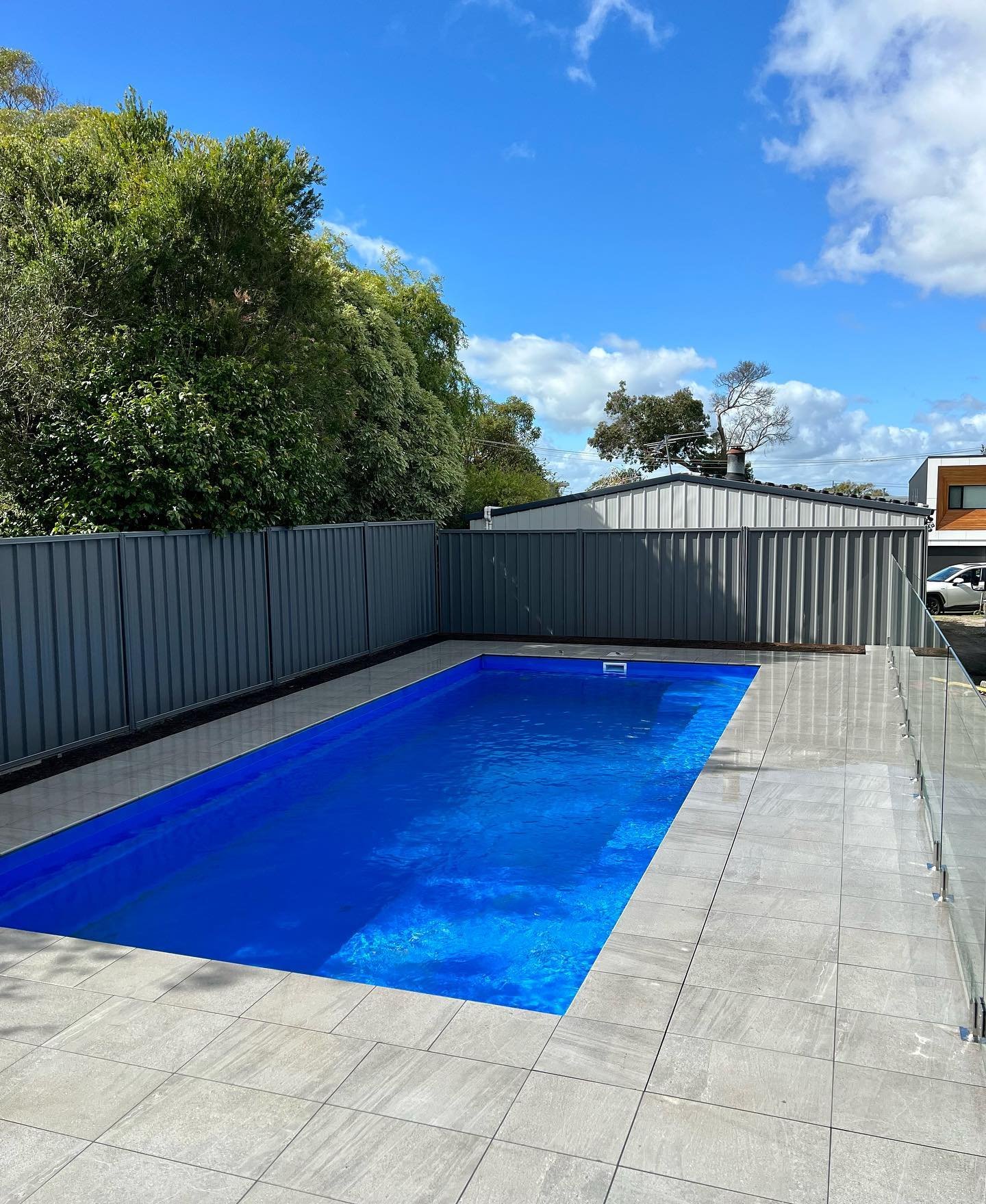 A rectangular above-ground swimming pool with blue water, surrounded by beige tiled patio, enclosed by a gray metal fence, with greenery and a building in the background under a blue sky with clouds.
