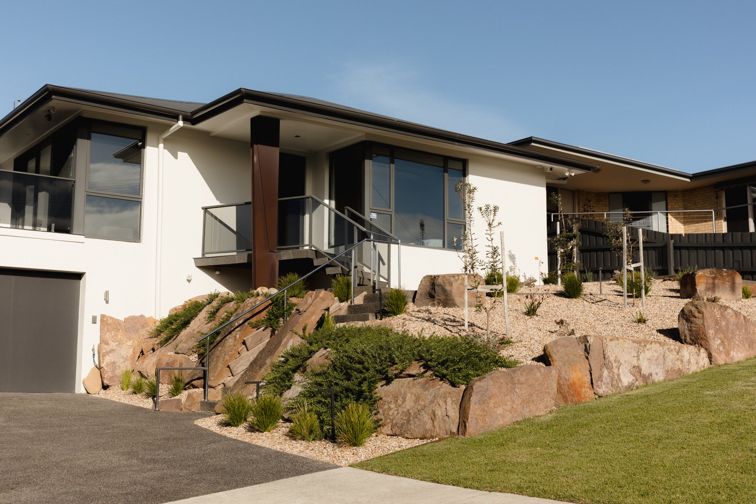 Modern house with white exterior, large windows, and a landscaped front yard with rocks, shrubs, and young trees.