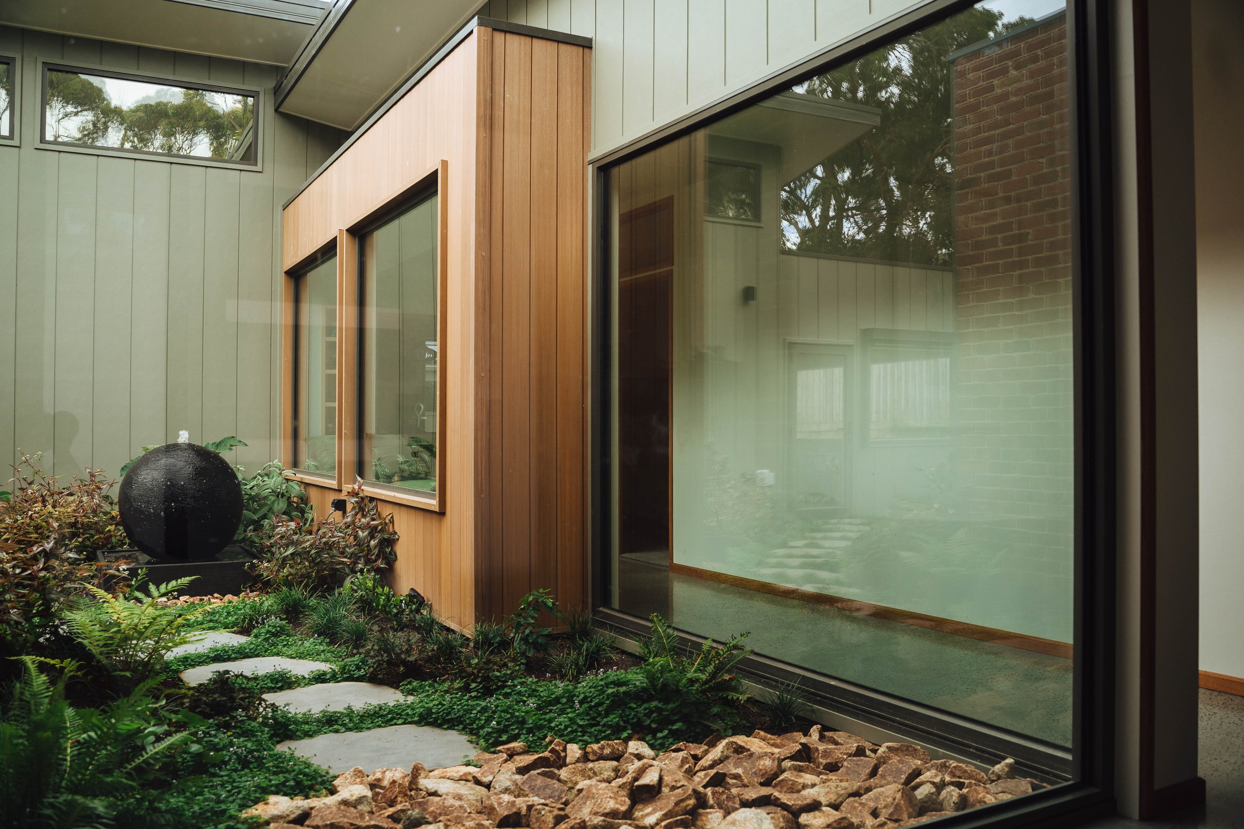 Modern house with large windows, wooden siding, and a landscaped garden with stepping stones, rocks, and a black spherical fountain.