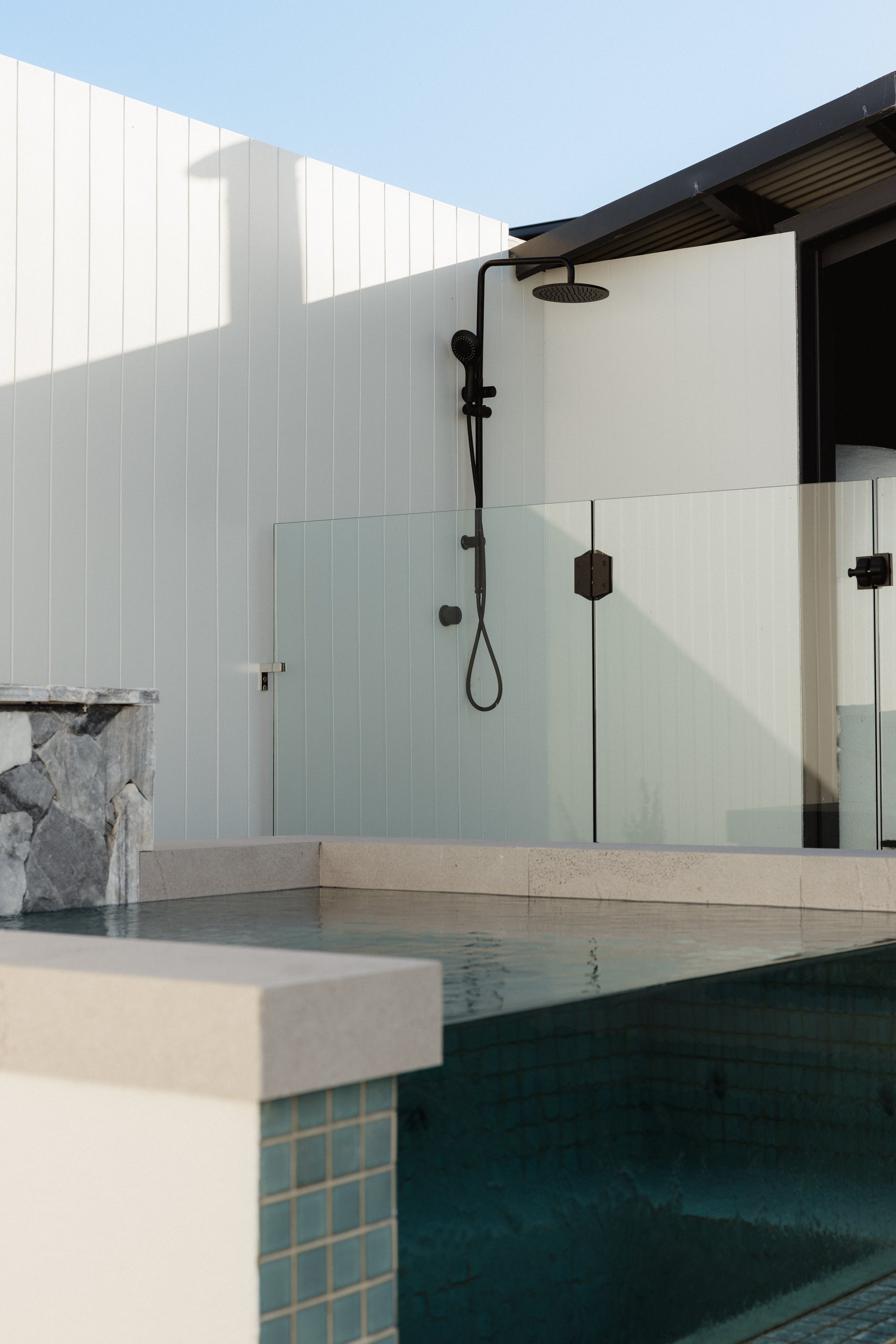 Outdoor shower area with black showerhead and white tiled walls, adjacent to swimming pool with stone and tile accents.