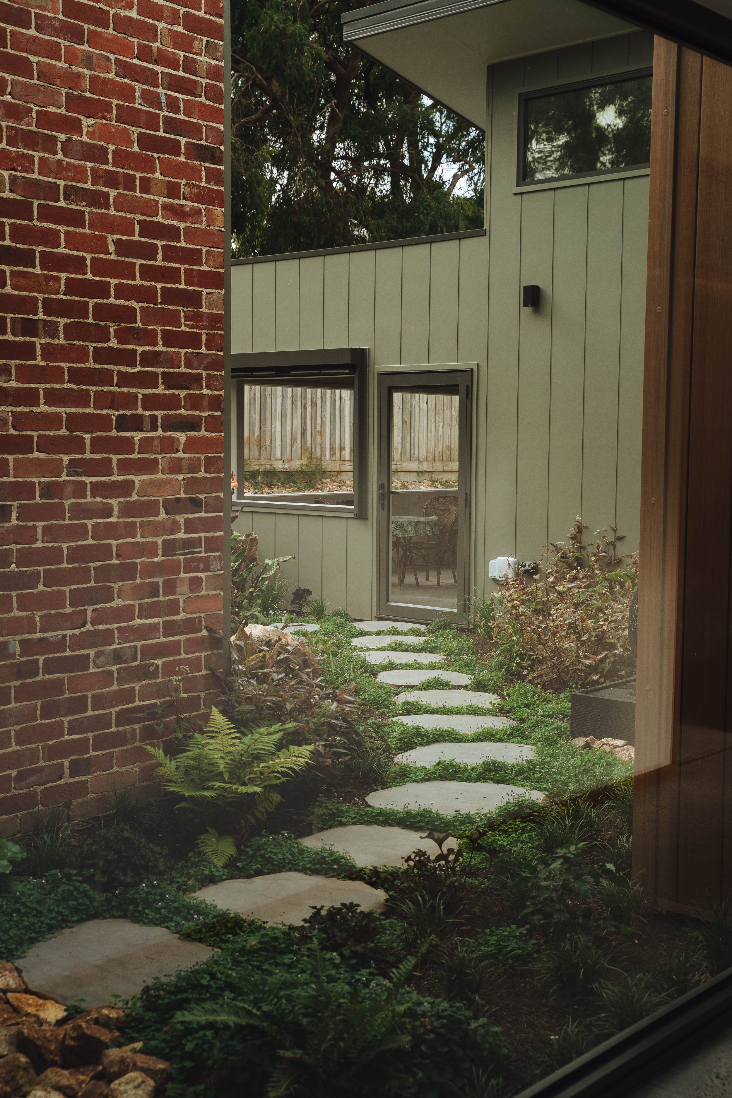 View through a window showing a backyard garden with a stone pathway, green plants, and modern patio furniture behind a glass door.