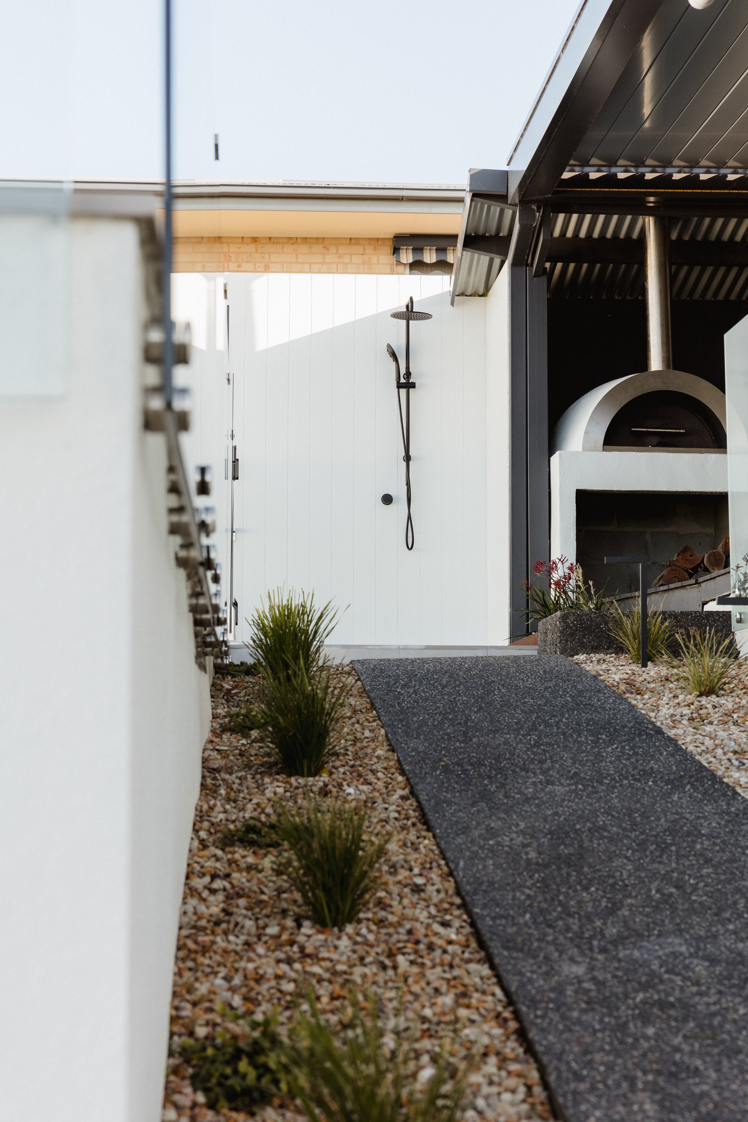 Outdoor kitchen and shower area with a black outdoor oven, plants in gravel bed, and a black pathway leading to a white door