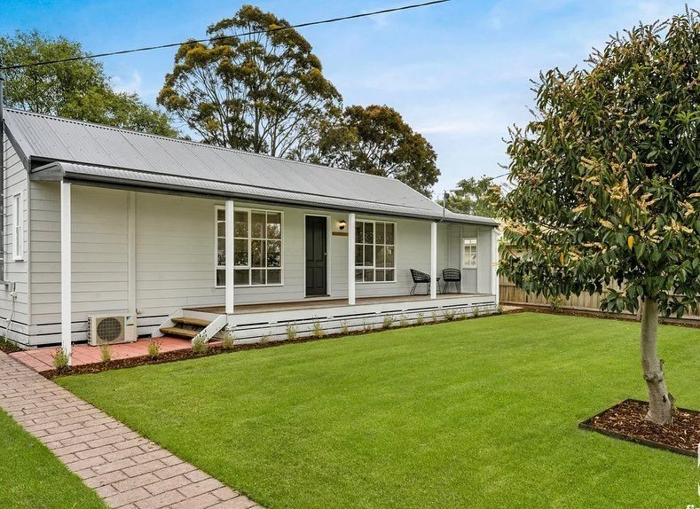 Front yard of a house with a green lawn, a brick pathway, a porch with two black chairs, a large tree, and a gray house with a metal roof.