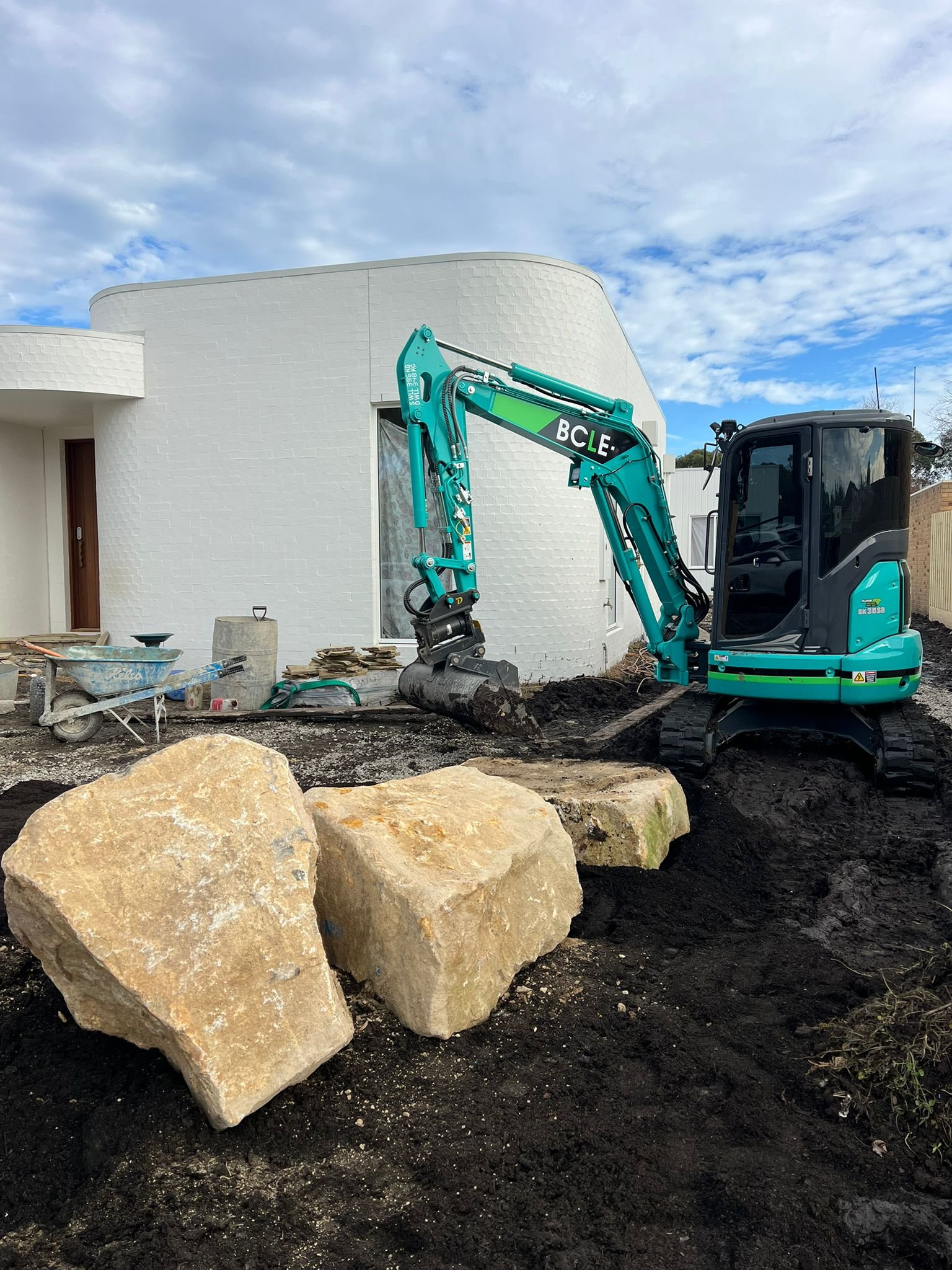 A small blue excavator is digging soil near a white modern building with rounded architecture, and large beige rocks are in the foreground.
