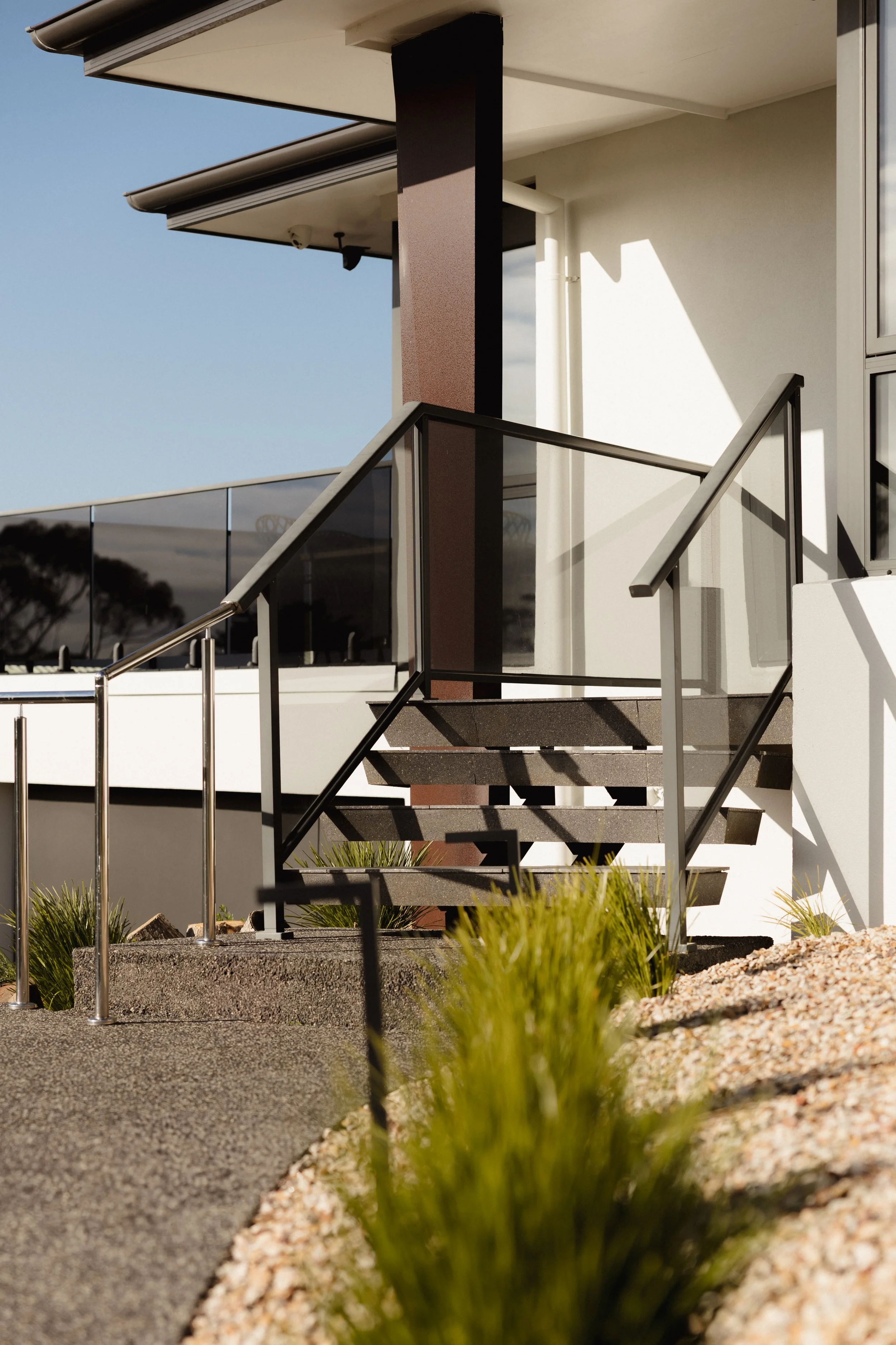 Exterior view of a modern building with a staircase, black railings, and brown column, with a blue sky and landscaped foreground.